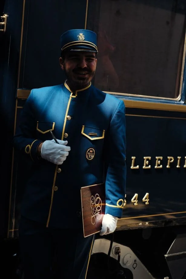 A man dressed as a vintage train conductor with a blue uniform and hat, holding a ticket and a booklet, standing in front of a train.