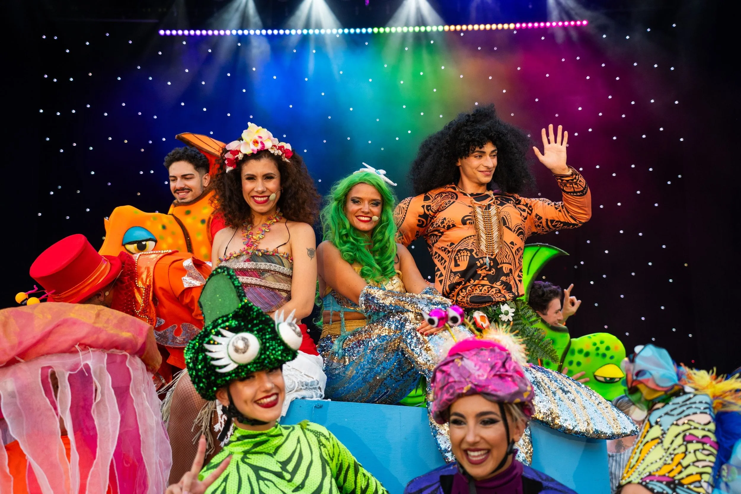 Group of people dressed in colorful, whimsical costumes, celebrating on a stage with rainbow-colored lighting and starry background.