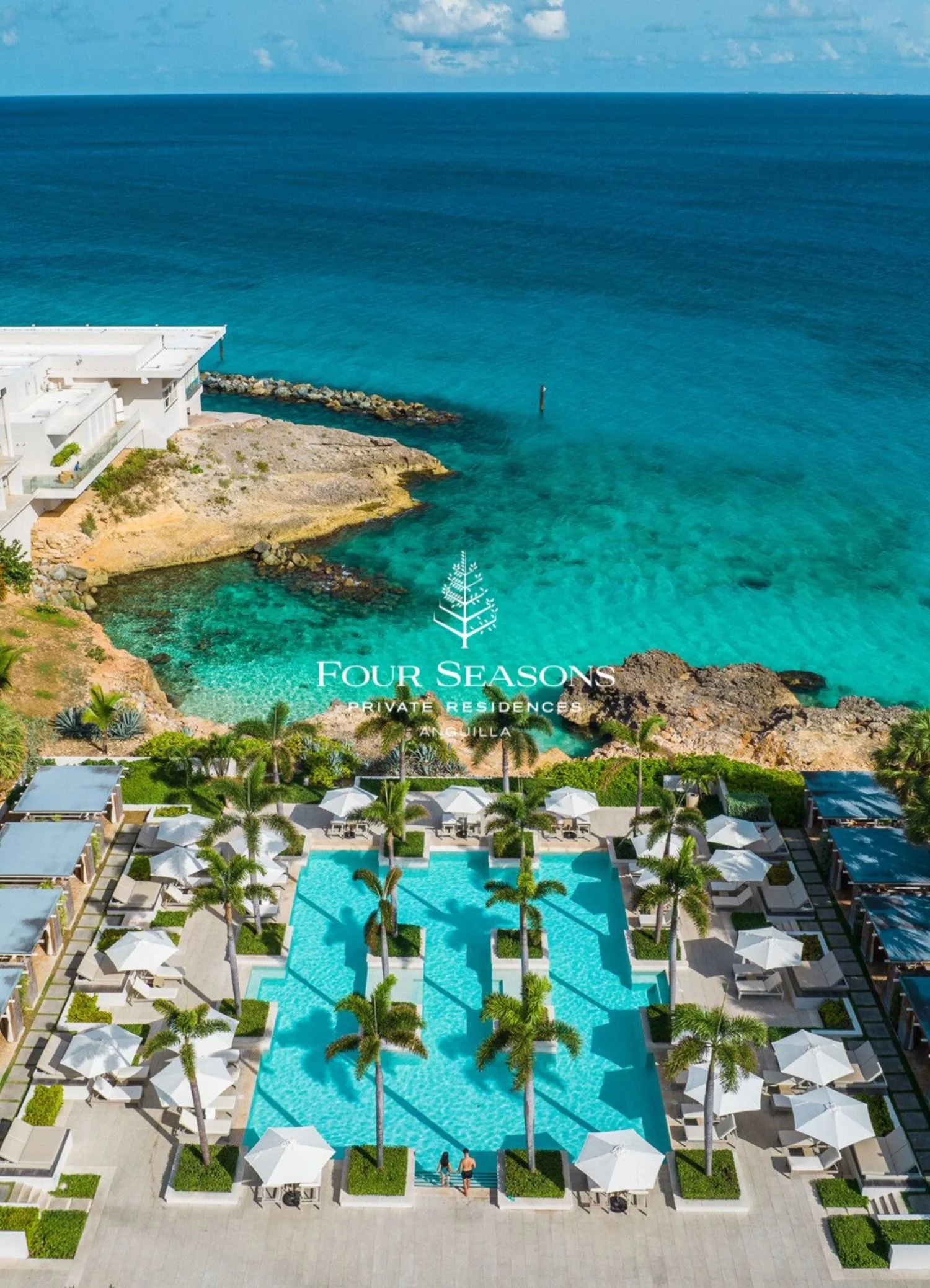Aerial view of a luxury resort with a large swimming pool, palm trees, white umbrellas, and lounge chairs, overlooking the ocean with a rocky pier and clear blue water.