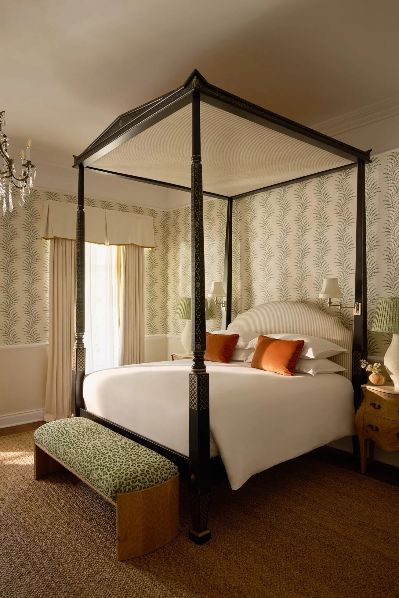 Elegant bedroom with a four-poster bed with black wood and decorative carvings. The bed has white bedding with orange pillows. A pair of wall-mounted lamps and a small wooden nightstand with a lamp are beside the bed. The room has patterned wallpaper, cream curtains, and a green upholstered bench at the foot of the bed. A chandelier is partially visible on the left.