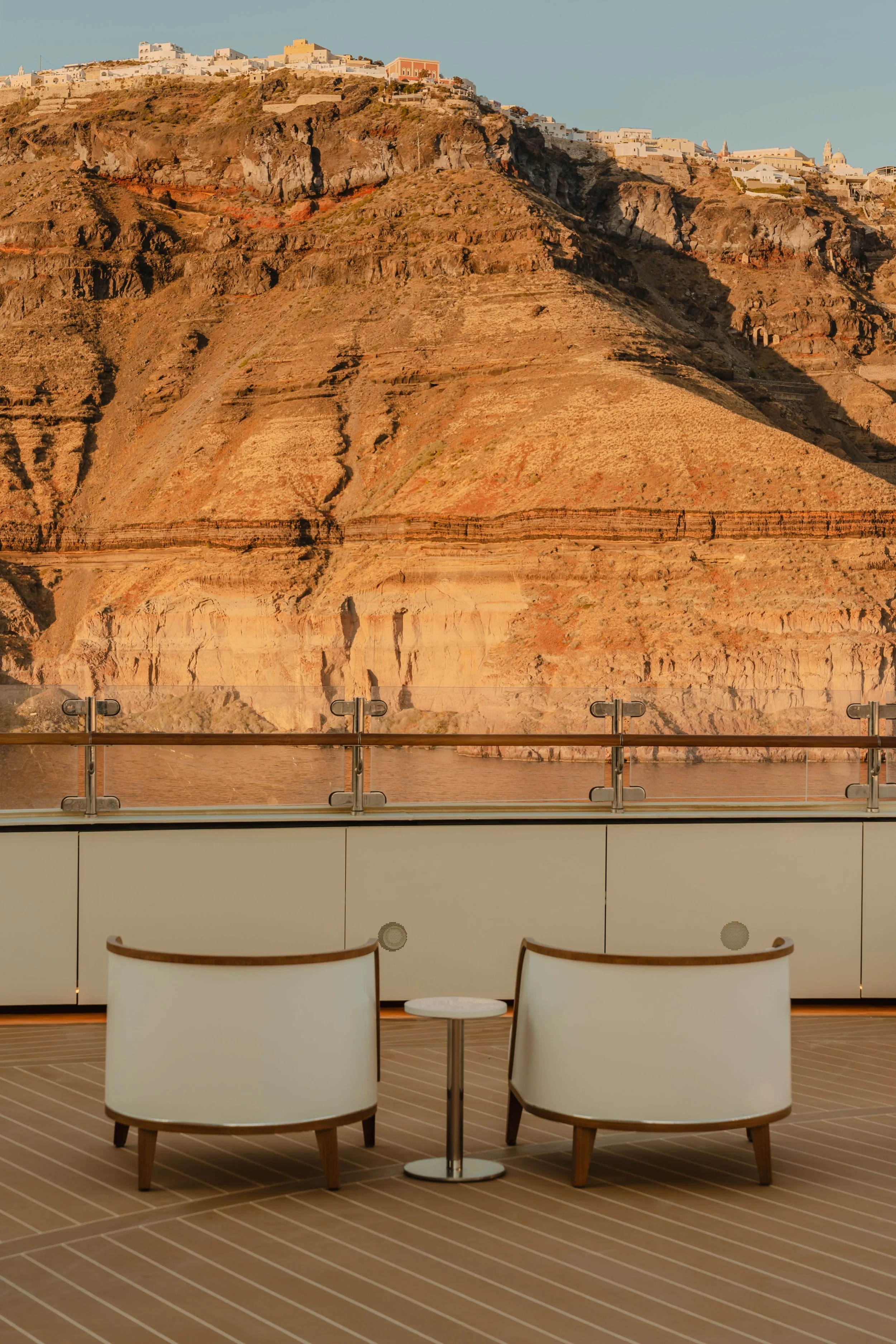 Interior of a cruise ship with two white chairs and a small round table in front, frente a a glass window showing a hillside with white buildings and rocky terrain.