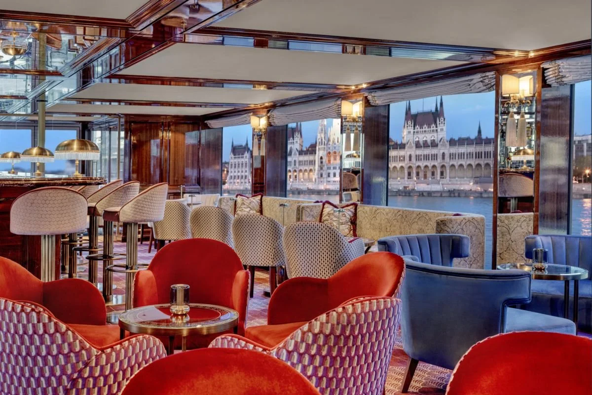 Interior of a bar or lounge with colorful chairs, large windows showing a cityscape with historic buildings and a river, and soft lighting.