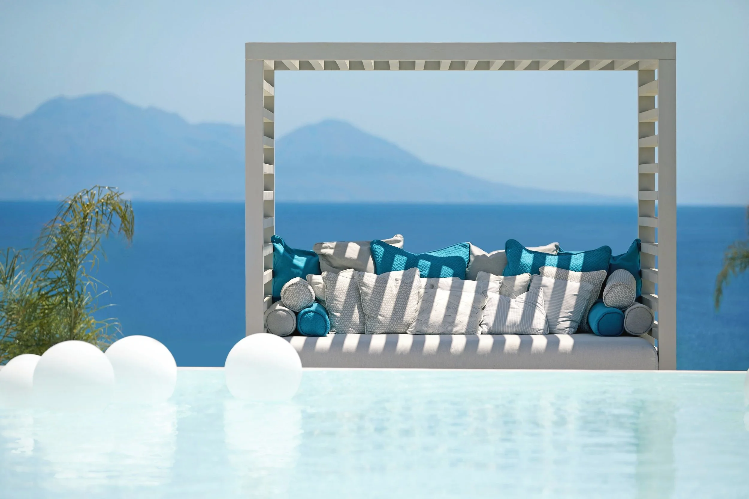 Outside seating area with a white wooden canopy frame, blue ocean in the background, and coastal mountains in the distance. The seating features a white and blue cushioned outdoor sofa with pillows.