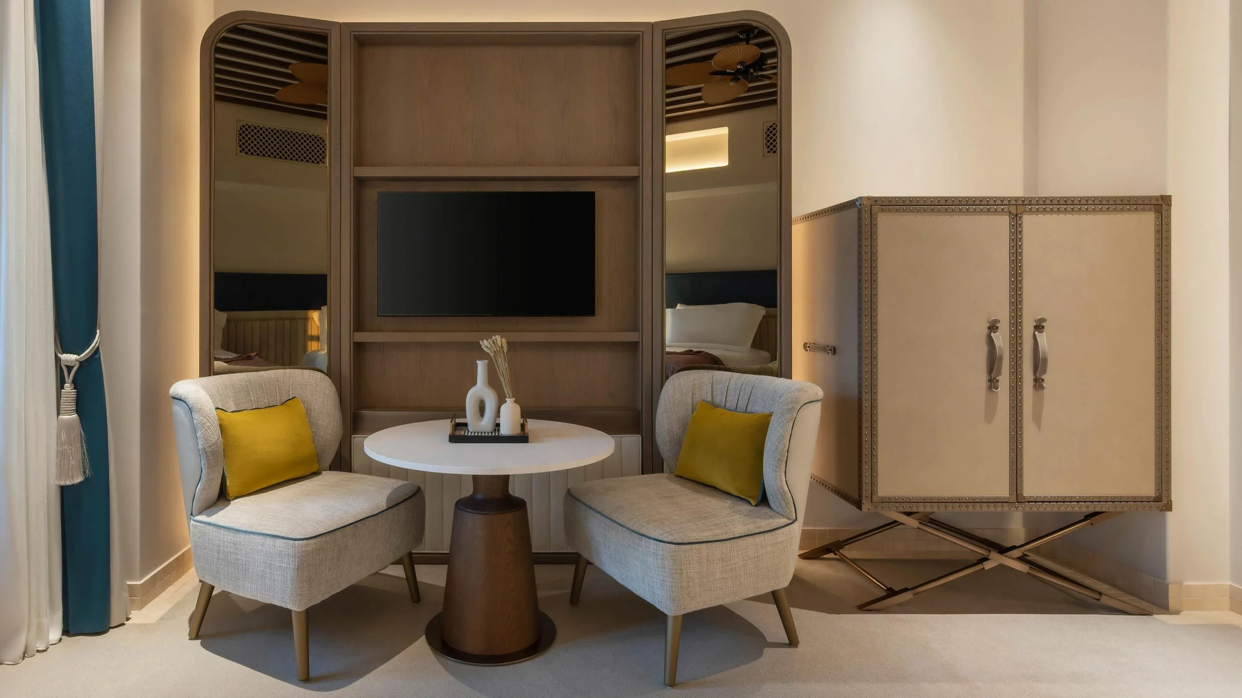 Living room with a round white table, two beige armchairs with yellow pillows, a decorative vase on the table, a wall with a mounted television, mirrored panels, a bed reflected in the mirrors, and a cabinet on the right