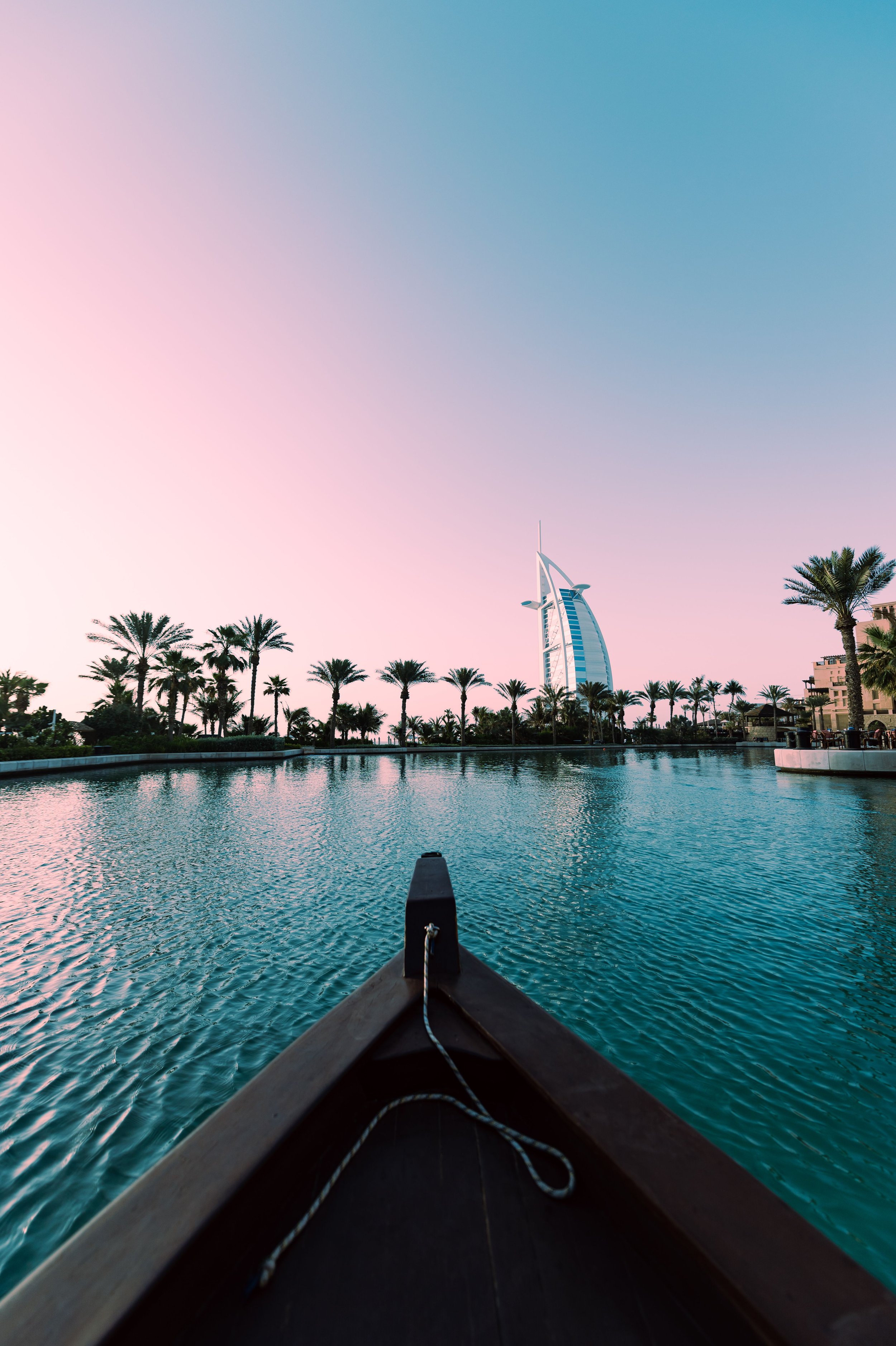 View from a boat on a calm body of water with a city skyline featuring the Burj Al Arab hotel in Dubai and palm trees along the shore at sunset.