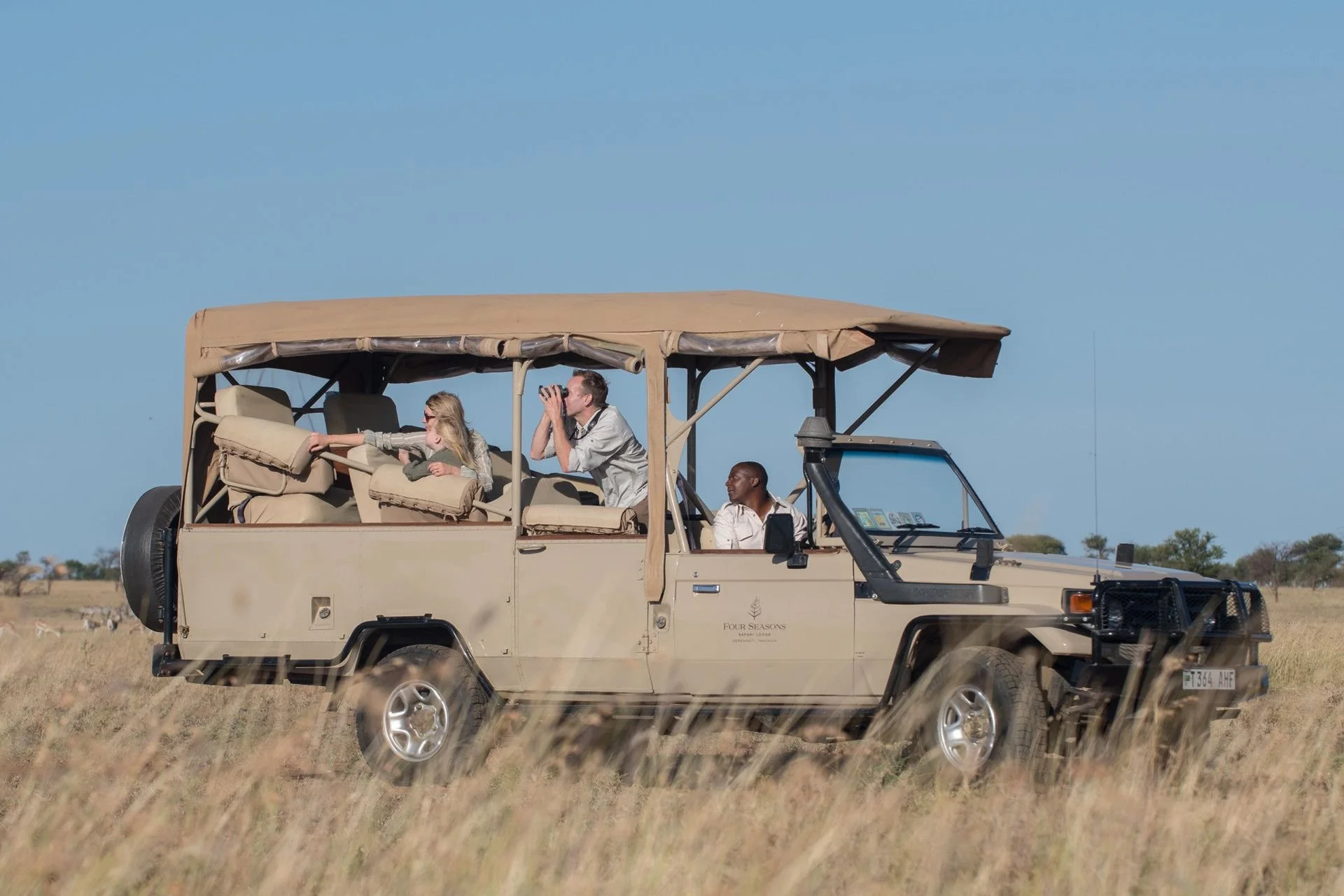 People riding in a safari vehicle across a grassy plain under a clear blue sky.