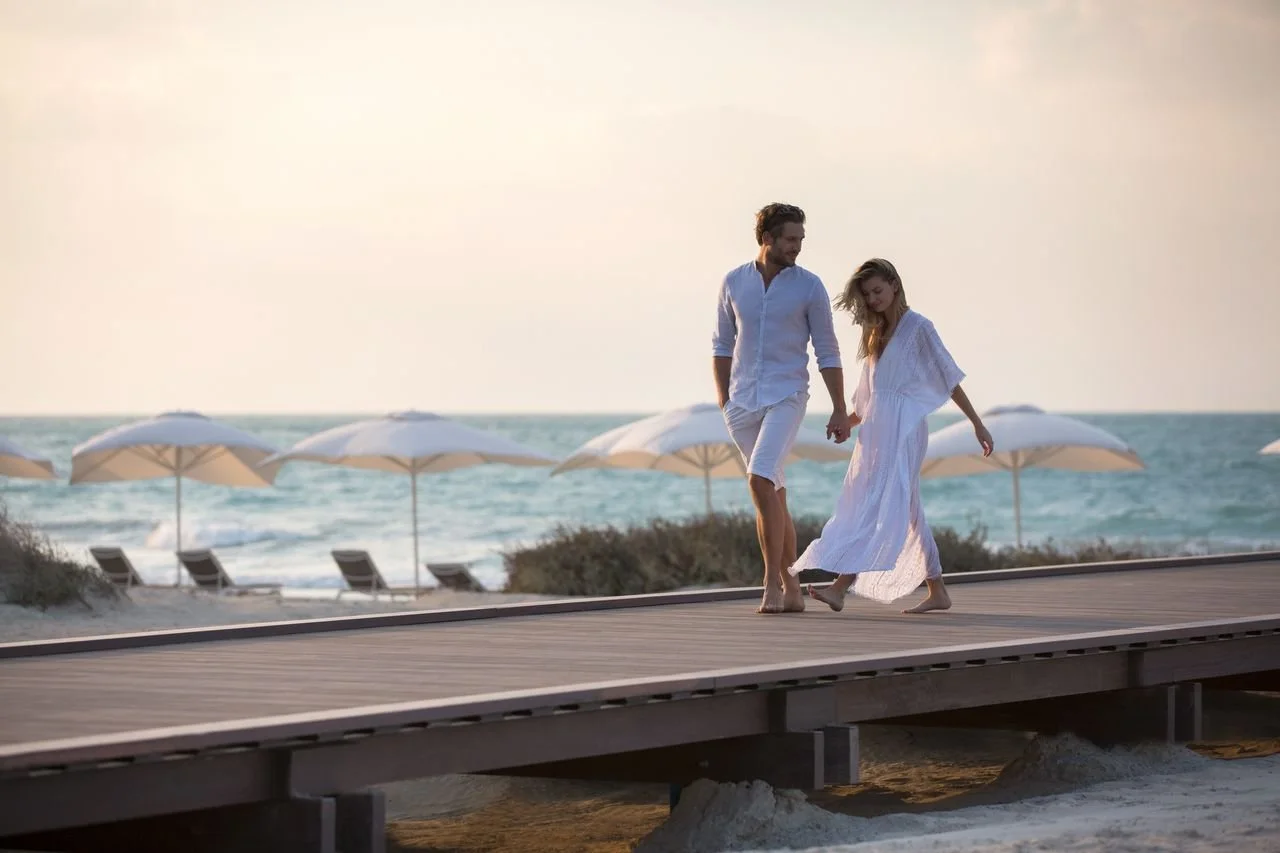 A couple walking hand in hand on a wooden boardwalk near the beach. The woman is wearing a long white dress, and the man is wearing a white shirt and shorts. Beach umbrellas and lounge chairs can be seen in the background, with the ocean and a pastel sky.