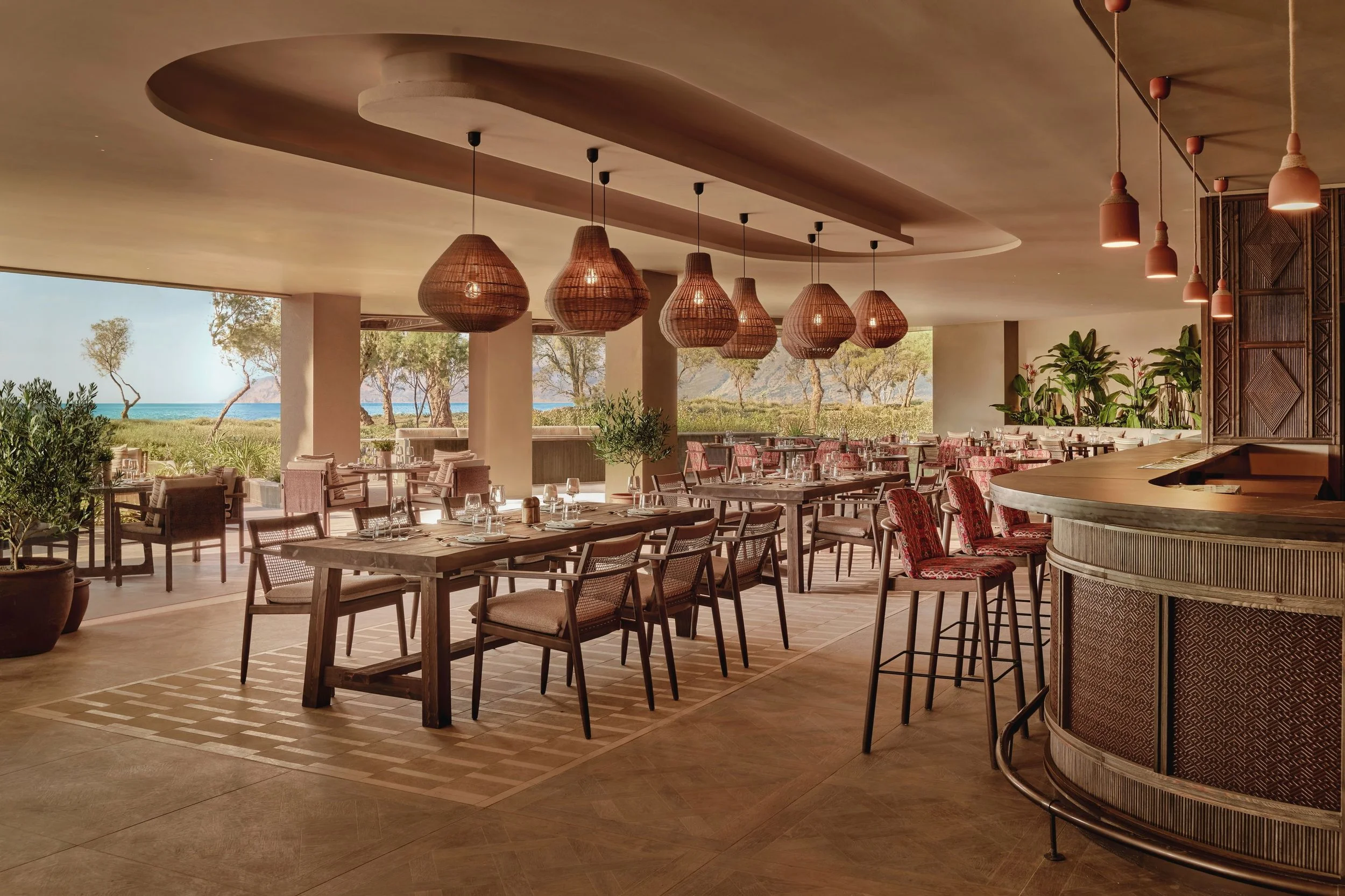 An open-air restaurant with a view of trees, grass, and the ocean, featuring wooden furniture, woven pendant lights, and potted plants.