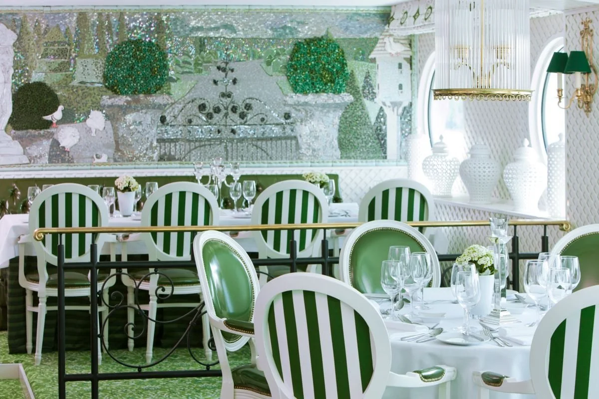 Elegant dining room with white and green striped chairs, white tablecloths, and wine glasses. Decor includes large white vases, green lamps, and a wall mural of a garden scene with flowers and greenery.