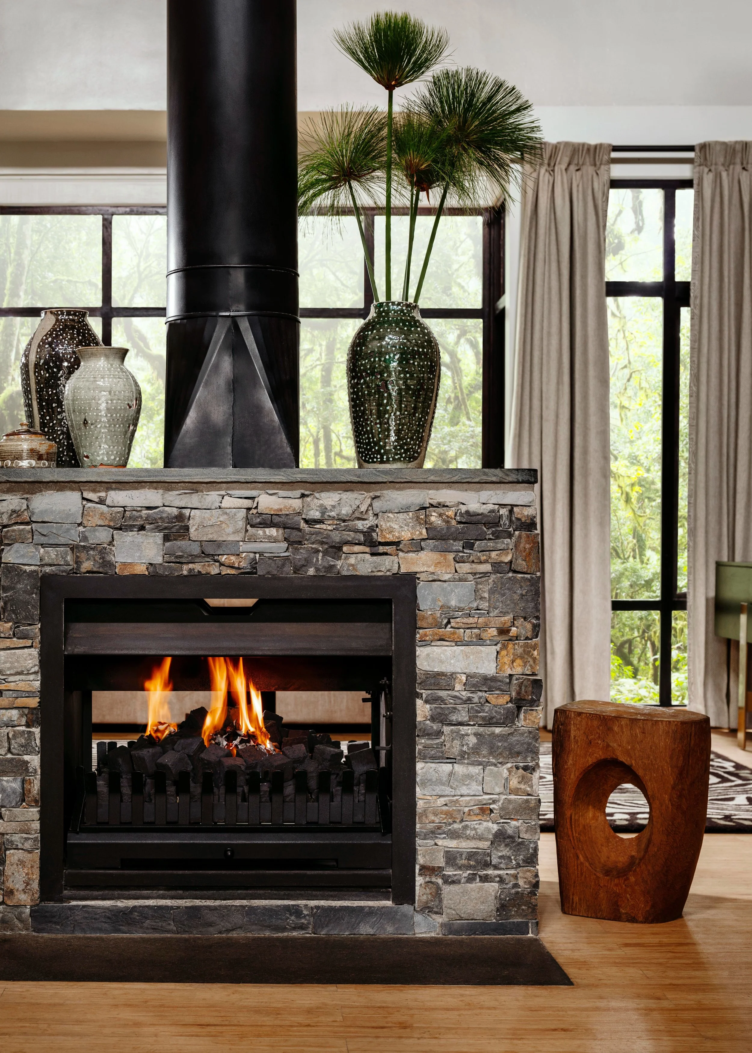 A cozy living room with a stone fireplace, lit with a fire. On top of the fireplace, there are decorative vases and a tall plant with long green leaves. There are large windows with beige curtains on either side, allowing natural light to enter. A wooden stool or sculpture is positioned next to the fireplace on the right side.