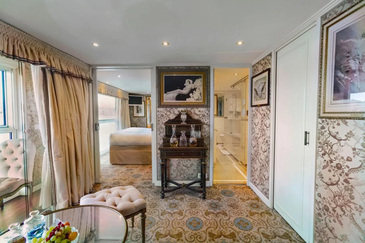 Elegant bedroom with floral wallpaper, cream curtains, tufted chair, glass table with fruit, and a bed in the background.
