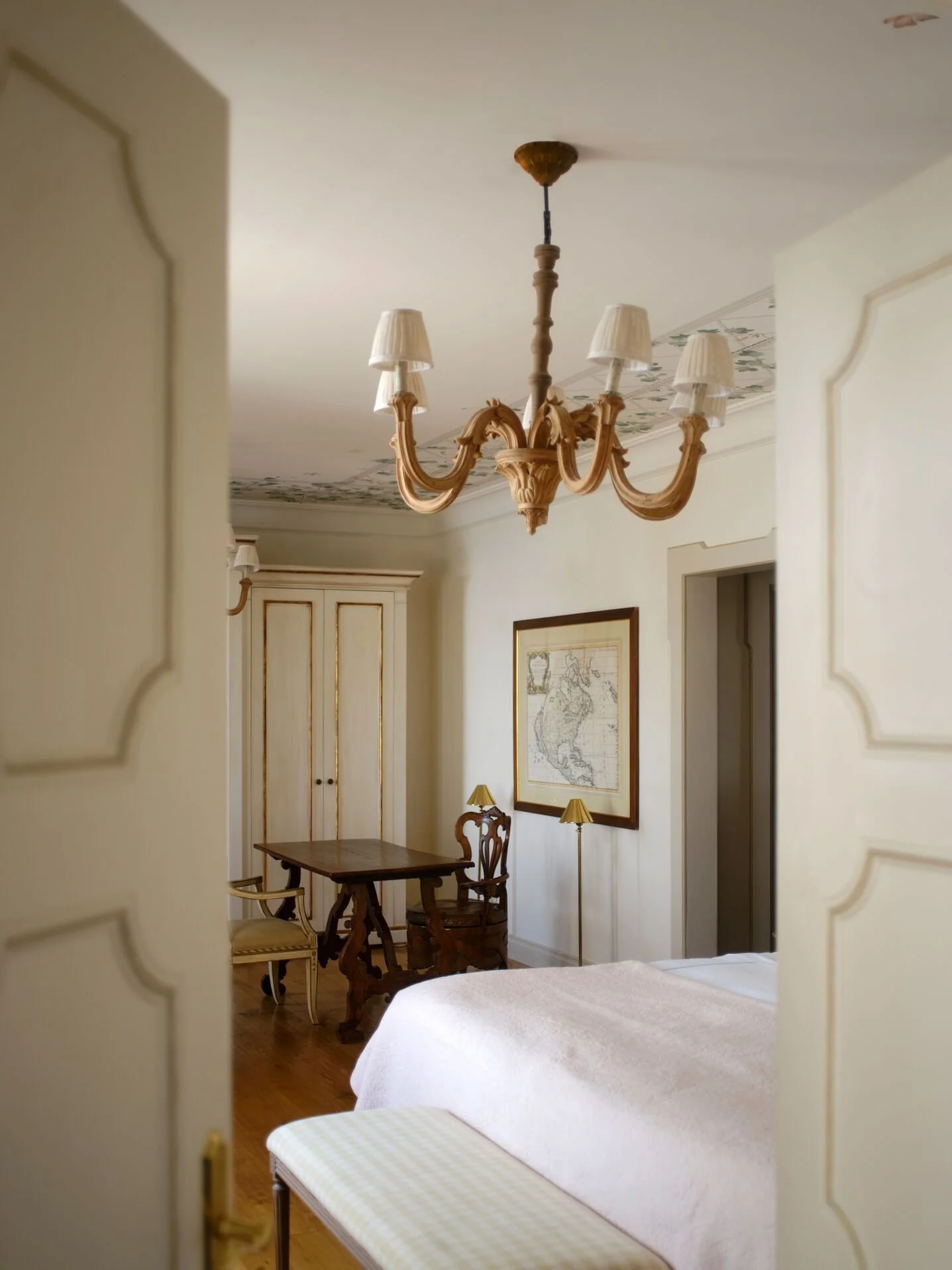 A dining room with a chandelier hanging from the ceiling, a table with chairs, a framed map on the wall, and table lamps.