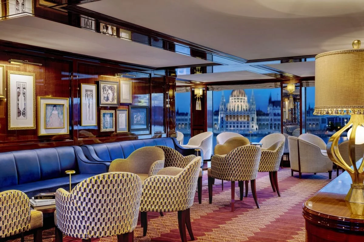 Elegant hotel lounge with upholstered chairs, a dark blue sofa, framed artwork on wood-paneled walls, a large window with a city skyline and historical building, and a gold lamp on a wooden table.