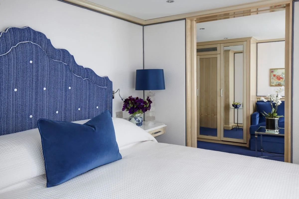 A neatly made hotel bed with a blue headboard, blue pillow, and white bedding in a room with a mirrored closet and blue armchair with a side table, inside a well-decorated hotel room.
