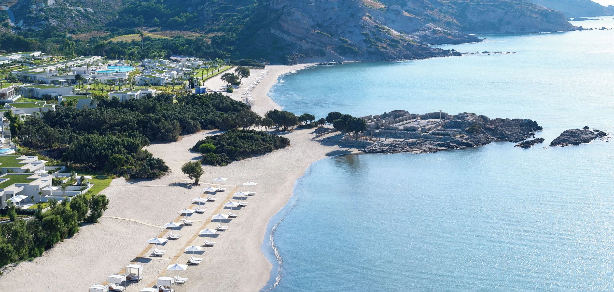 Aerial view of a coastal resort with white buildings, a sandy beach with sunbeds and umbrellas, green trees and shrubs, rocky outcroppings, and calm blue ocean water, with hills in the background.