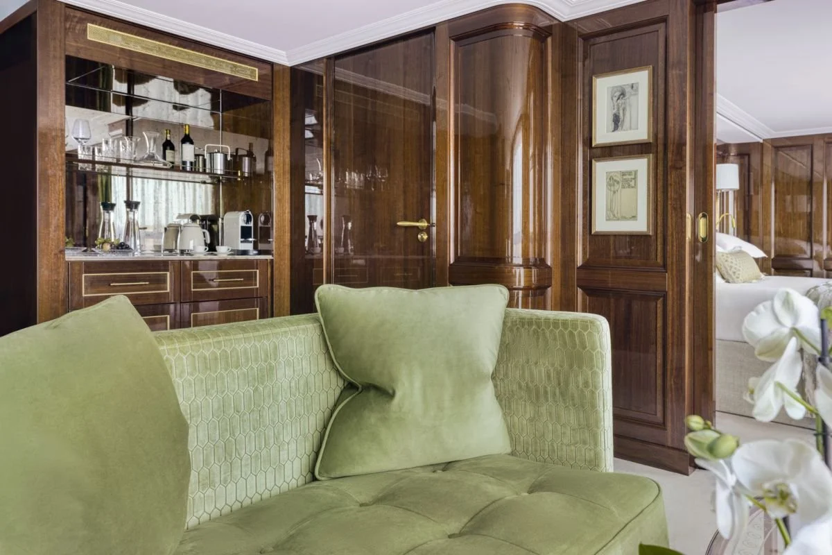 A hotel suite living room with a green sofa, wooden paneled walls, and a bedroom visible through an open doorway.