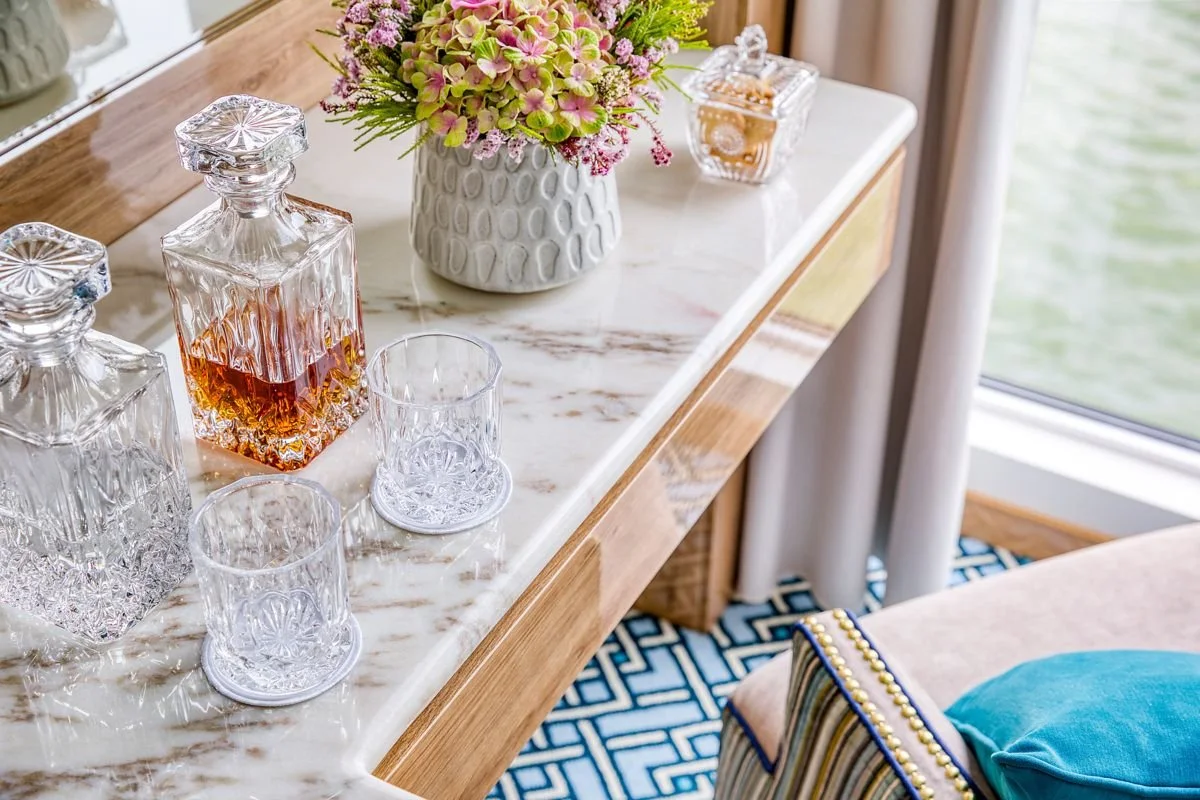 A marble-topped side table with crystal decanters, glasses, a vase of pink and green hydrangeas, and a bowl of snacks near a window with white curtains.
