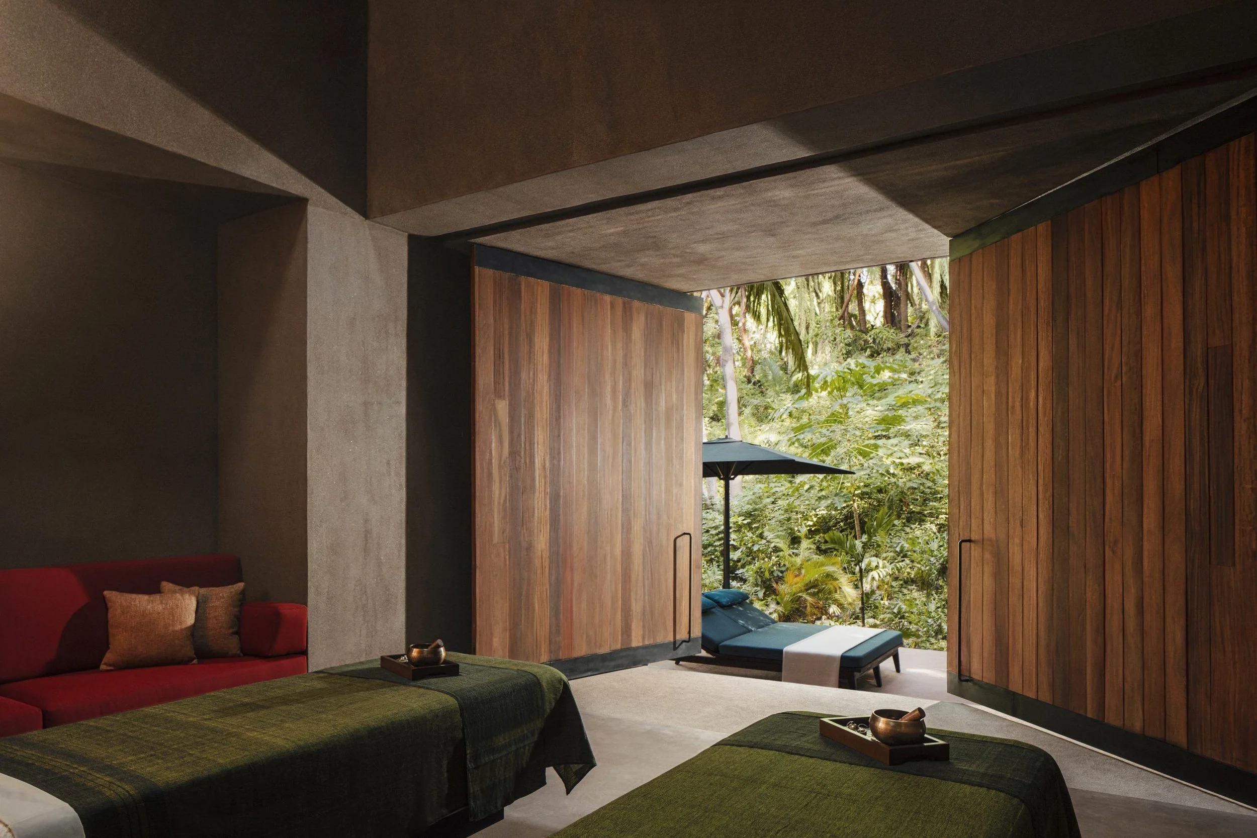 Modern interior room with large window opening to a lush green outdoor landscape, featuring wooden and concrete walls, a red sofa with pillows, and low tables with decorative objects.