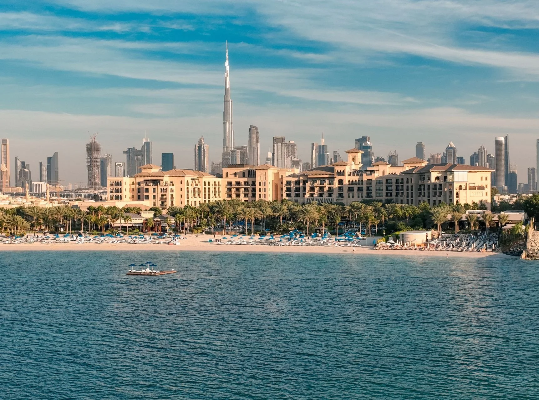 A city skyline with tall skyscrapers, including the Burj Khalifa, overlooking a beach with lounge chairs and palm trees. A boat with a blue umbrella is on the water.
