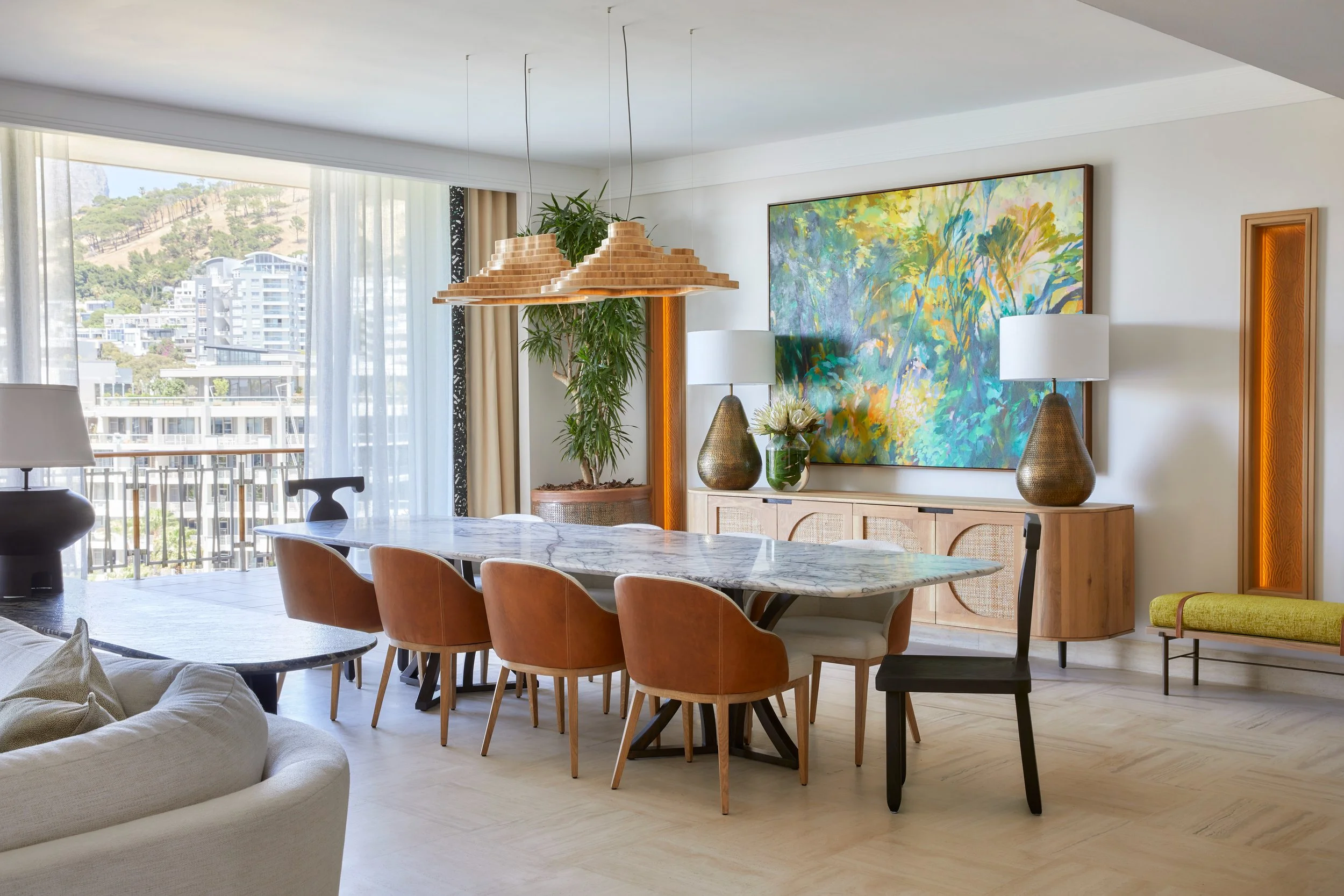 Living room with a marble dining table, brown chairs, a white sofa, artwork on the wall, two table lamps, a large plant, and a window with a city view.