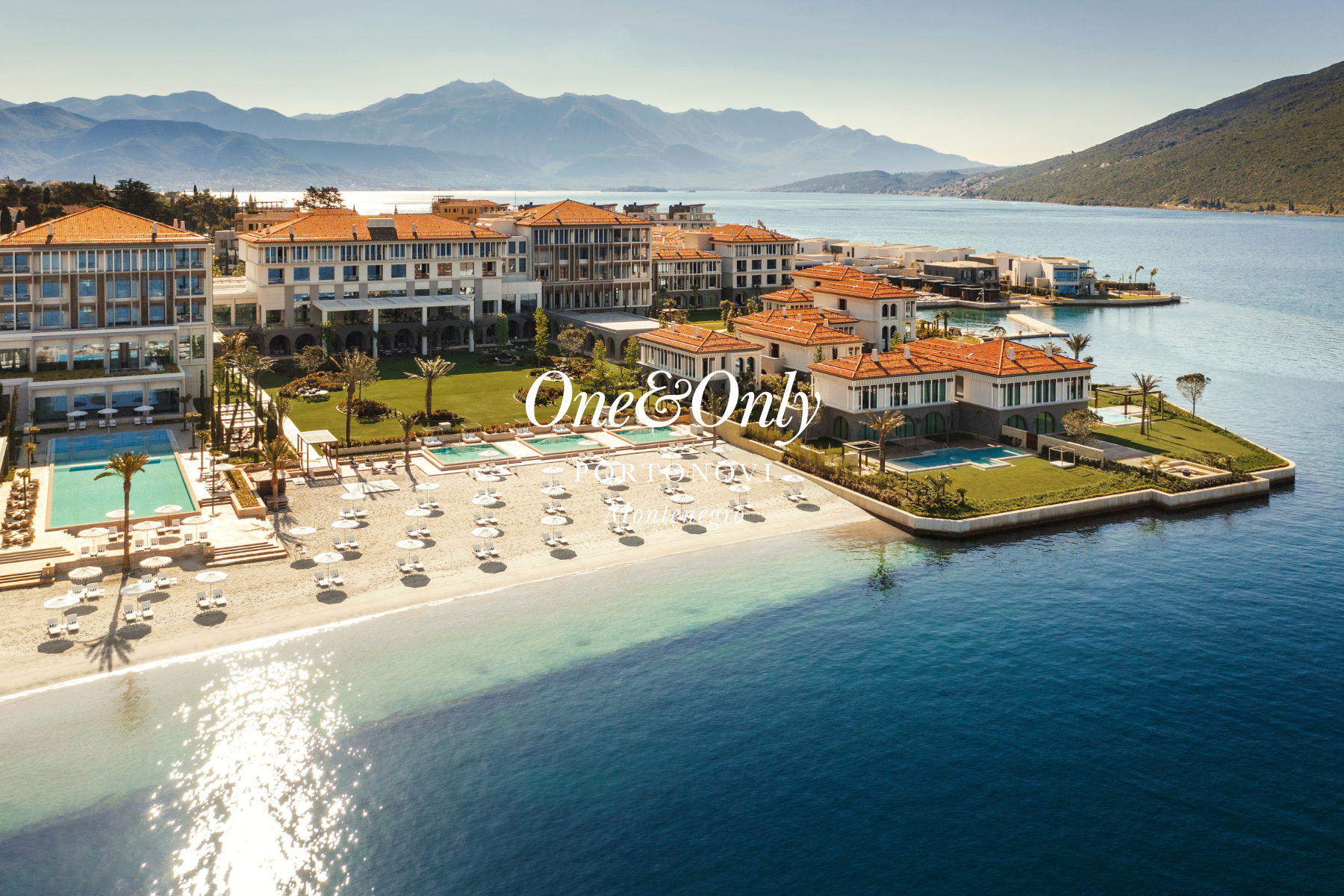 An aerial view of a luxurious beachfront resort in Porto Novi, Montenegro, with multiple pools, landscaped gardens, modern buildings, and a calm bay with mountains in the background.