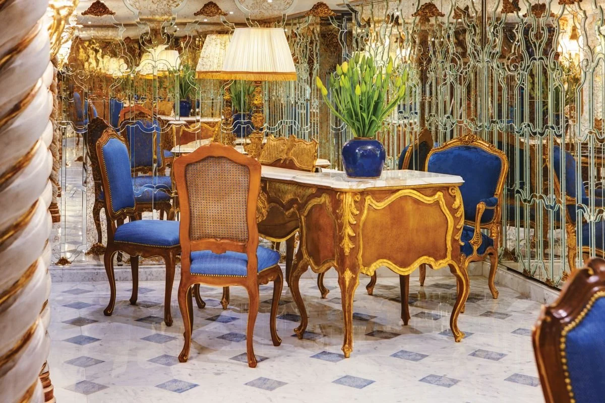 Elegant dining area with ornate gold-trimmed wooden chairs with blue upholstery, a marble-topped table, a blue vase with yellow flowers, decorative mirrors, and a lamp in a luxurious setting.