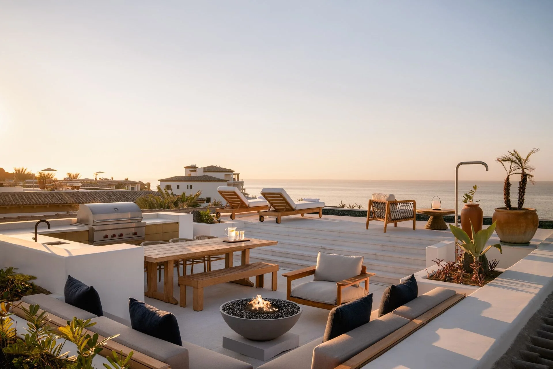An outdoor rooftop patio with lounge chairs, a fire pit, potted plants, a barbecue grill, a dining table with benches, and a view of the ocean at sunset.