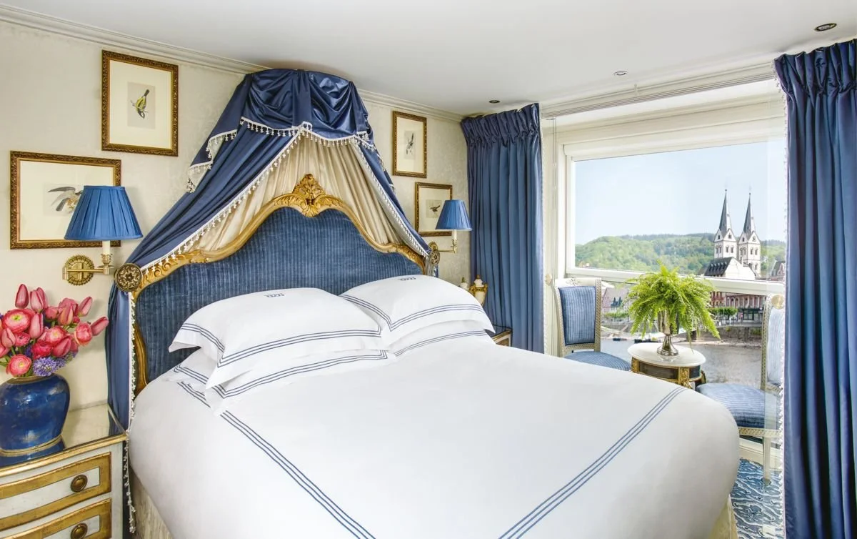 A luxurious bedroom with a large bed featuring a blue and gold ornate headboard and matching canopy, blue curtains, a window with a view of a church steeple and green hills, two blue lamps, wall art of birds, a side table with a vase of pink flowers, and a small table with a green plant.