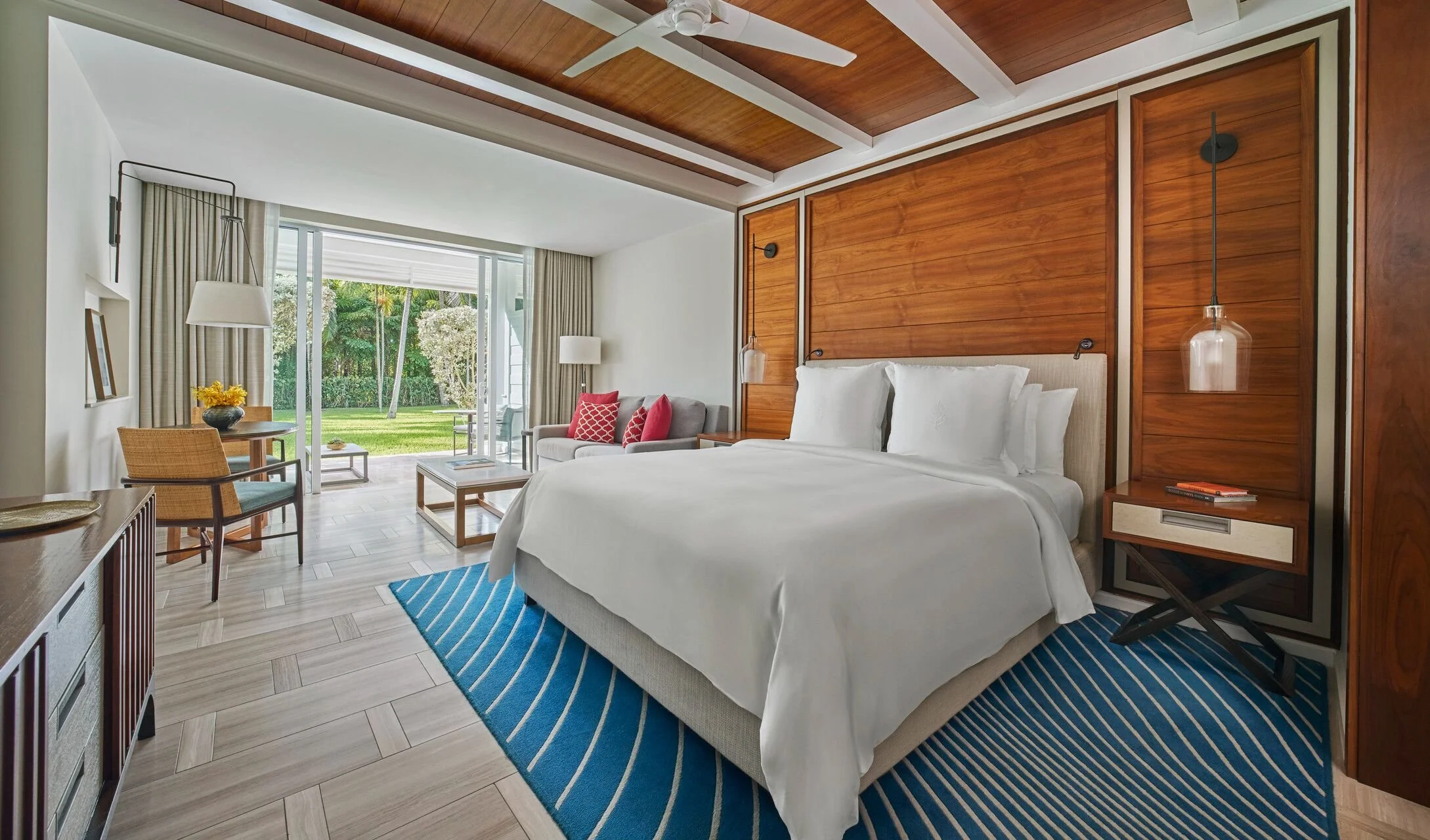 Modern hotel room with a large bed, wooden accent wall, and sliding glass door leading to a green outdoor space. The room has a ceiling fan, bedside lamps, and a seating area with a table and chairs.