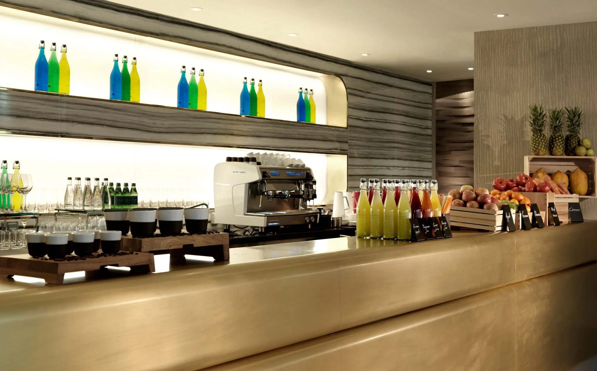 A modern bar counter with various bottles, glasses, and an espresso machine, set against a marble wall with a shelf of colorful bottles and fresh pineapples and fruits on a side shelf.
