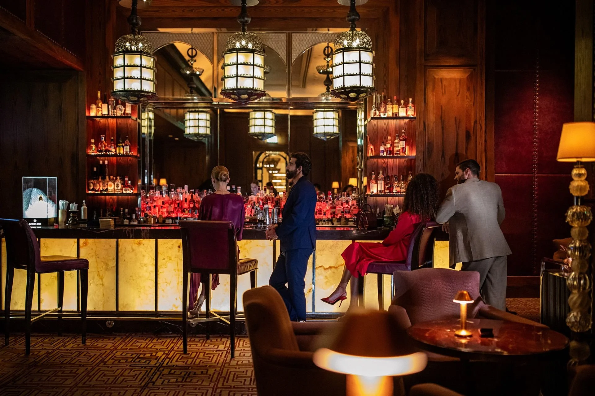 Interior of a upscale bar or lounge with warm lighting, wood paneling, and a marble bar counter. Several people are socializing, including a man in a blue suit and a woman in a red dress. The bar is decorated with various bottles of alcohol and illuminated by hanging lamps with intricate designs.