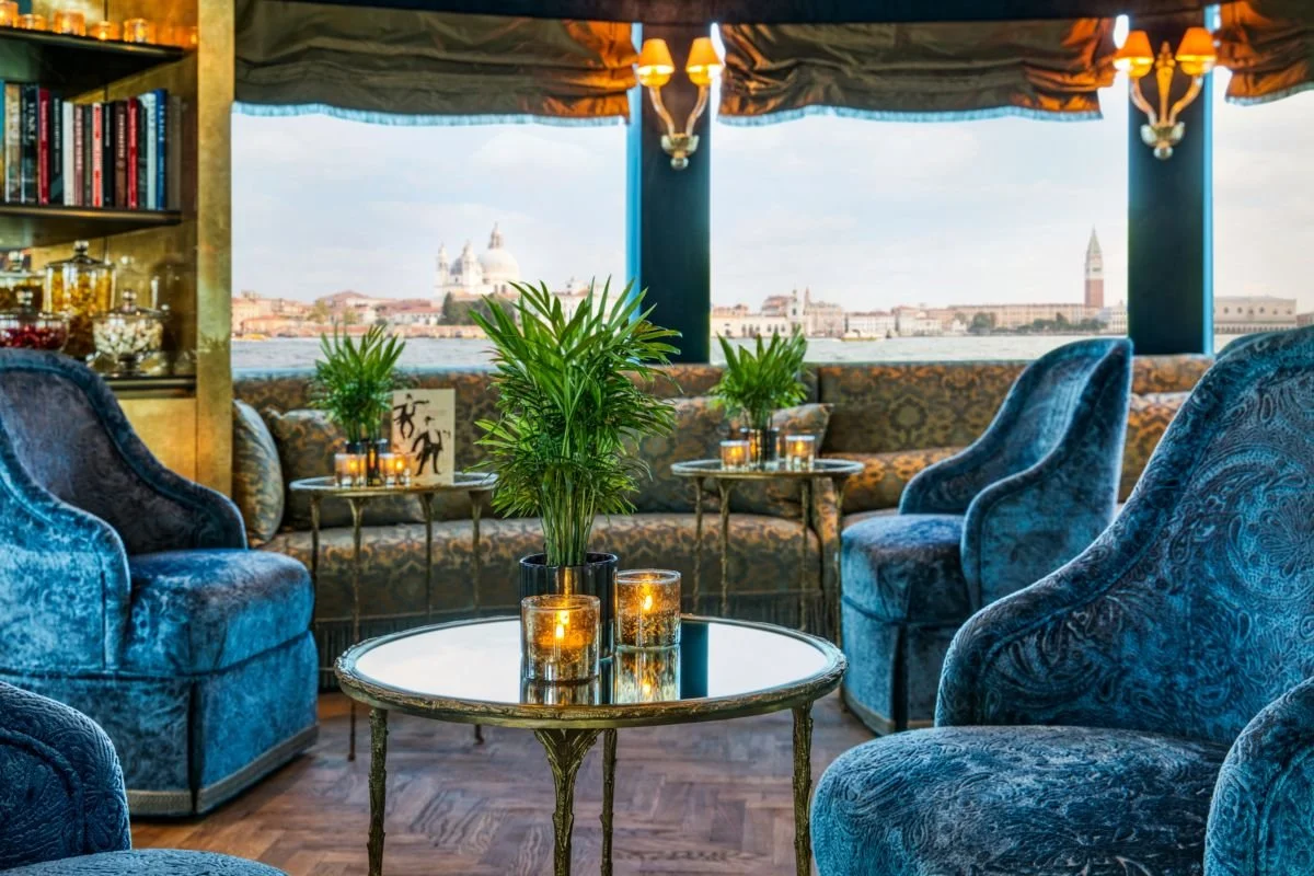 Elegant lounge with plush blue velvet armchairs, glass-topped tables, and lush green plants, overlooking a scenic waterfront view with historic buildings in the distance.