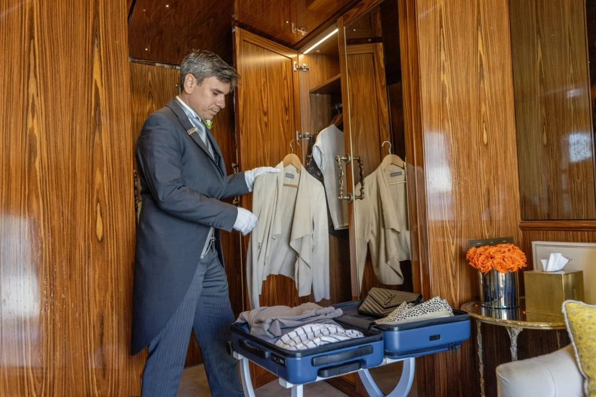 A man in a suit packing clothes into a suitcase inside a wooden-paneled room, with an open wardrobe and a side table with orange flowers.