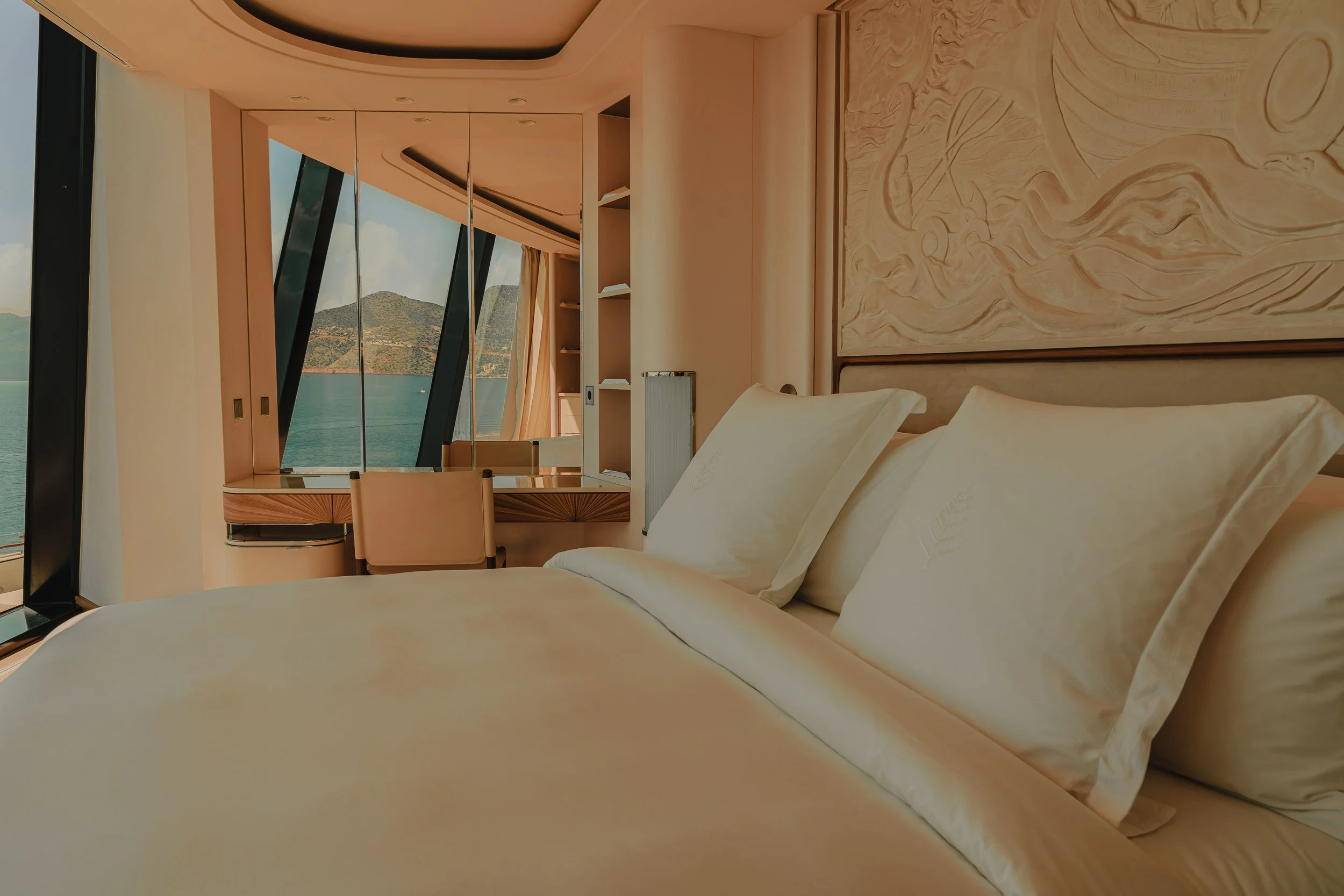 A hotel room with a bed, pillows, a wall with textured artwork, a large window showing a view of mountains and water, and a desk with a chair.