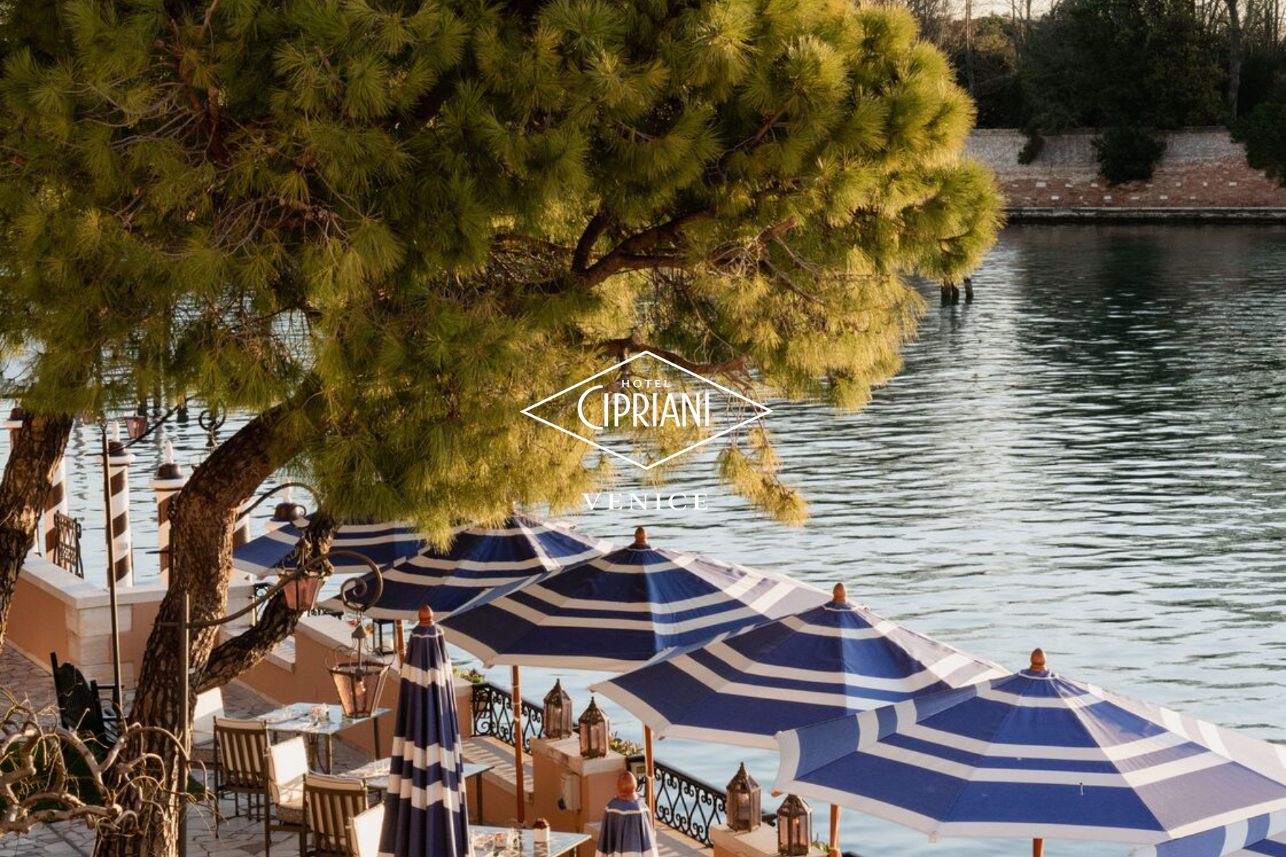 Outdoor dining area with tables and chairs under blue and white striped umbrellas along a waterfront with a large green tree in the foreground and water in the background in Venice.