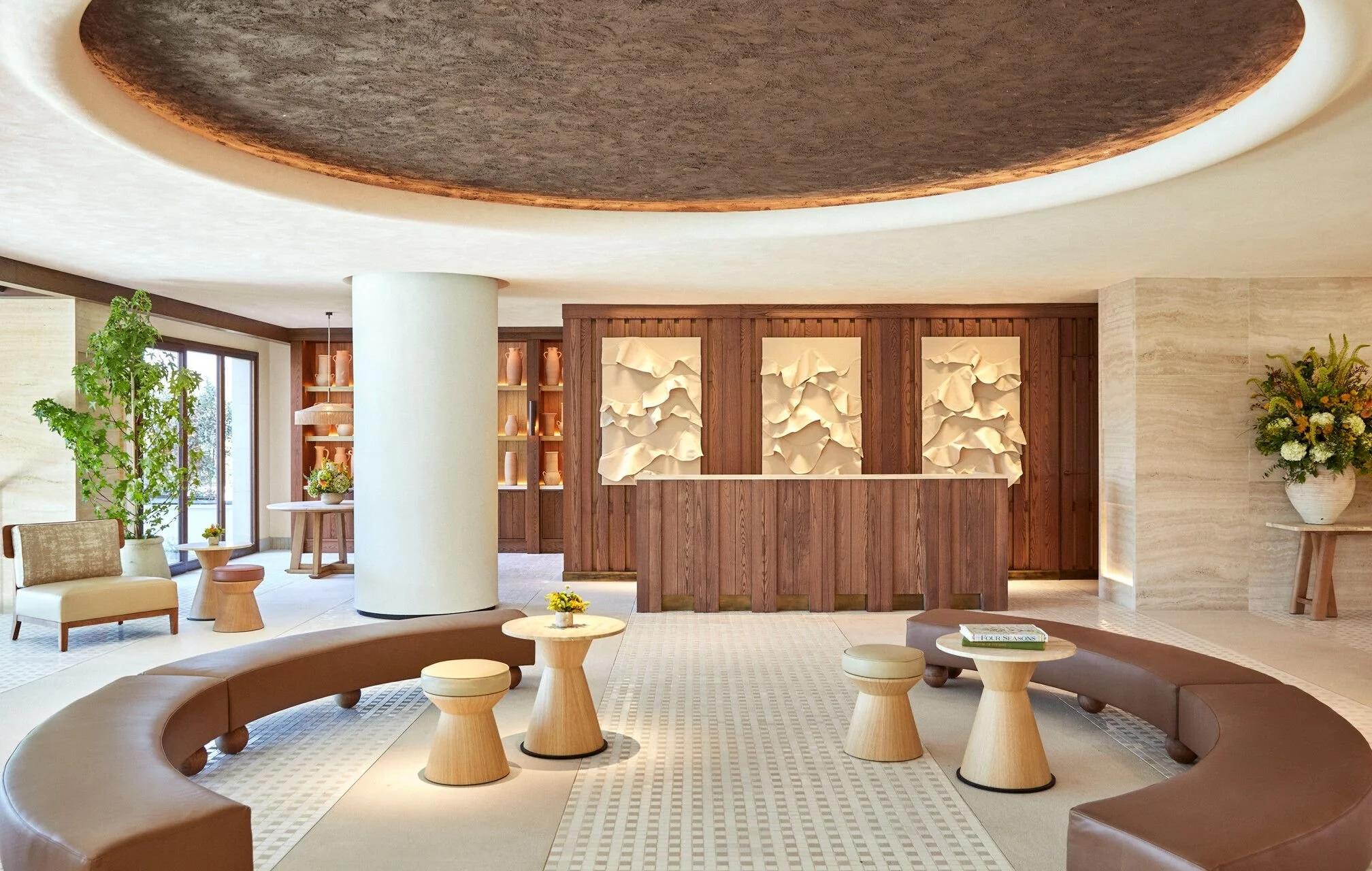 Interior of a modern hotel lobby featuring a large circular brown leather sofa, small round wooden tables, a wooden reception desk, decorative wall art, and lush green plants.