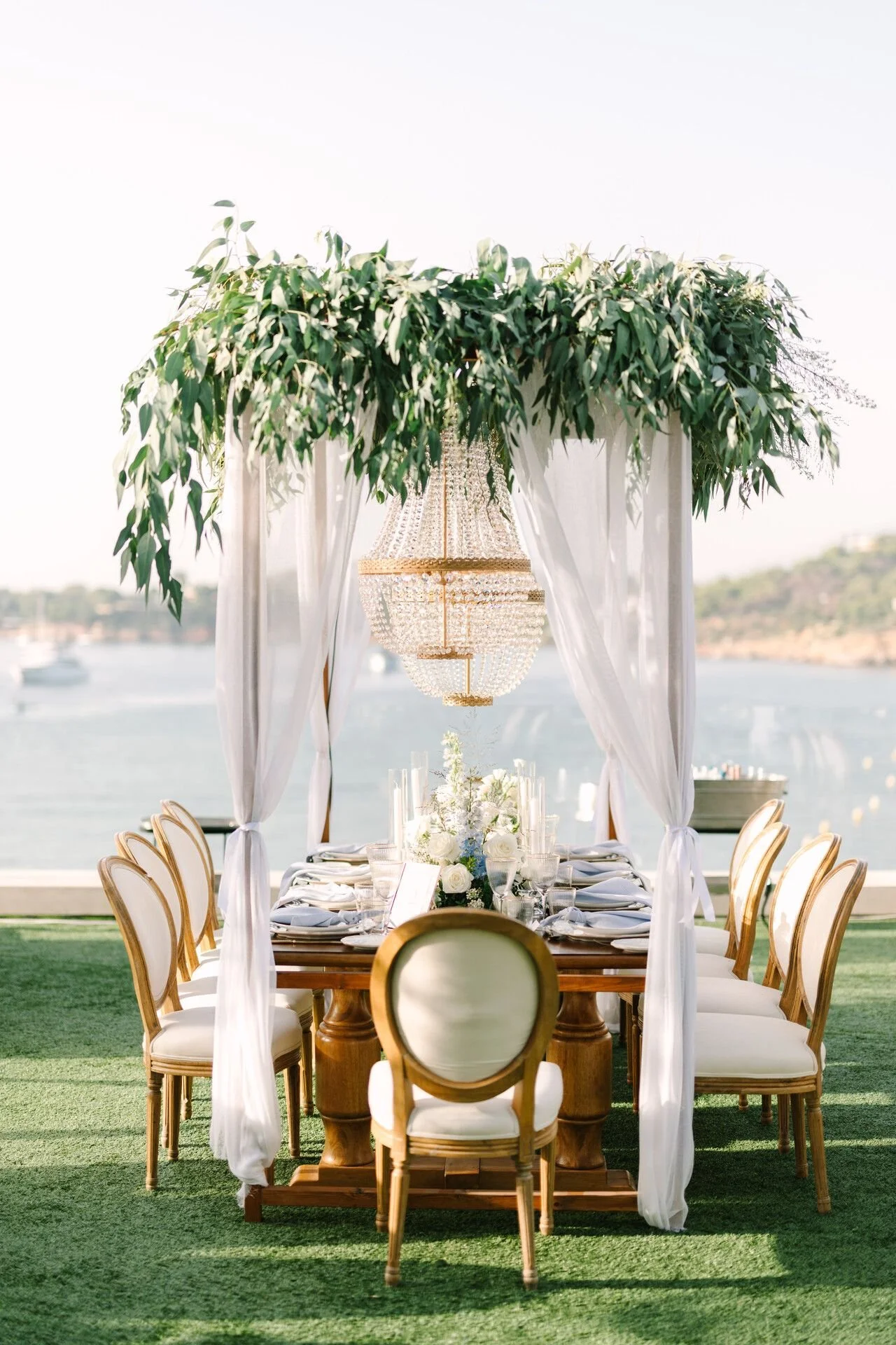 Elegant outdoor dining setup with a long table, white chairs with gold accents, and a canopy adorned with greenery and a chandelier, overlooking water and distant hills.
