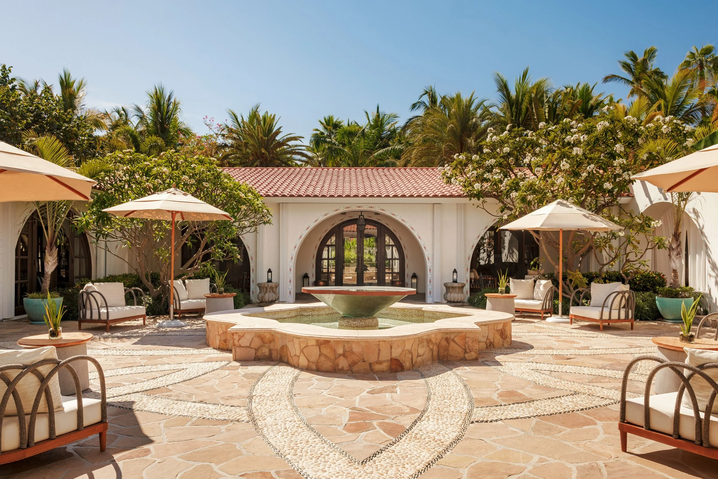 A luxurious outdoor patio with a stone fountain in the center, surrounded by cushioned chairs, potted plants, and patio umbrellas, with lush green palm trees and flowering trees in the background under a clear blue sky.