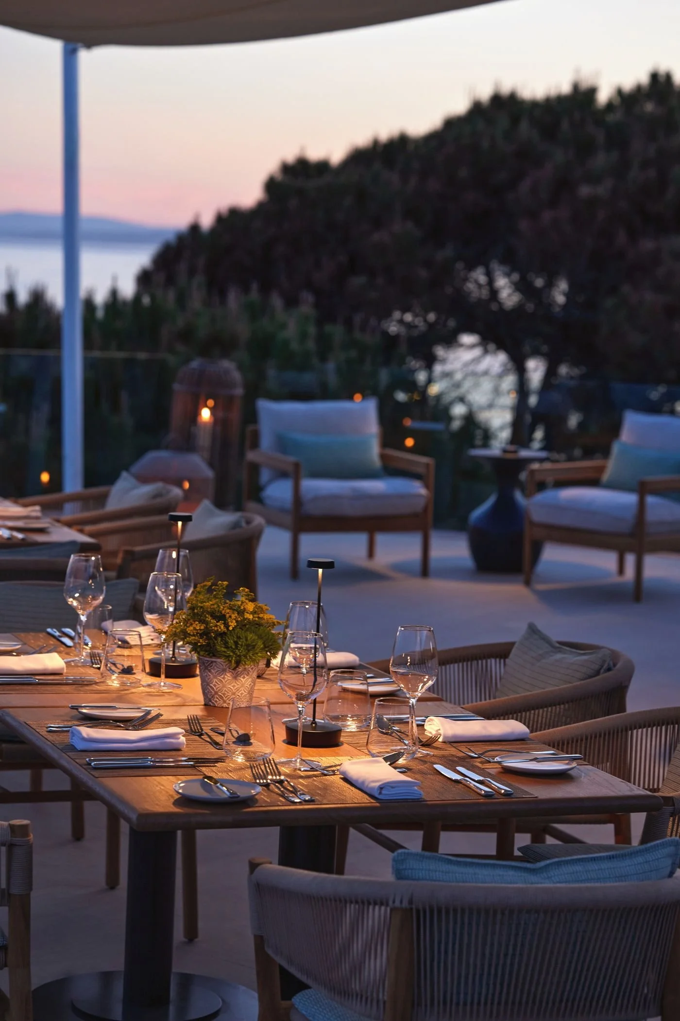 An outdoor dining area with a wooden table set with wine glasses, plates, silverware, and a small potted plant, illuminated softly with candlelight. In the background, there are cushioned chairs and lounge seating, a large tree, and a scenic view of a body of water and distant hills at dusk.