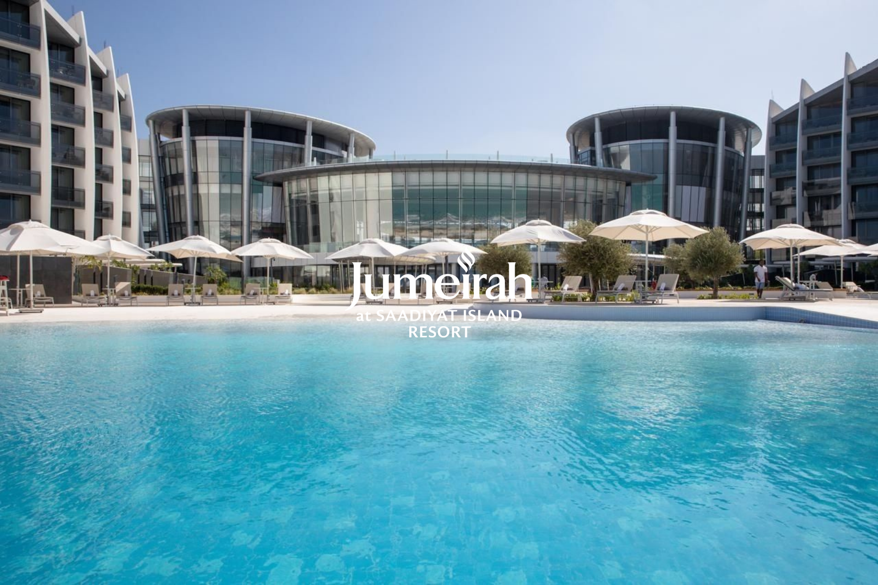 Luxury resort with a swimming pool, glass building, lounge chairs, umbrellas, and trees, at Jumeirah at Saadiyat Island Resort.