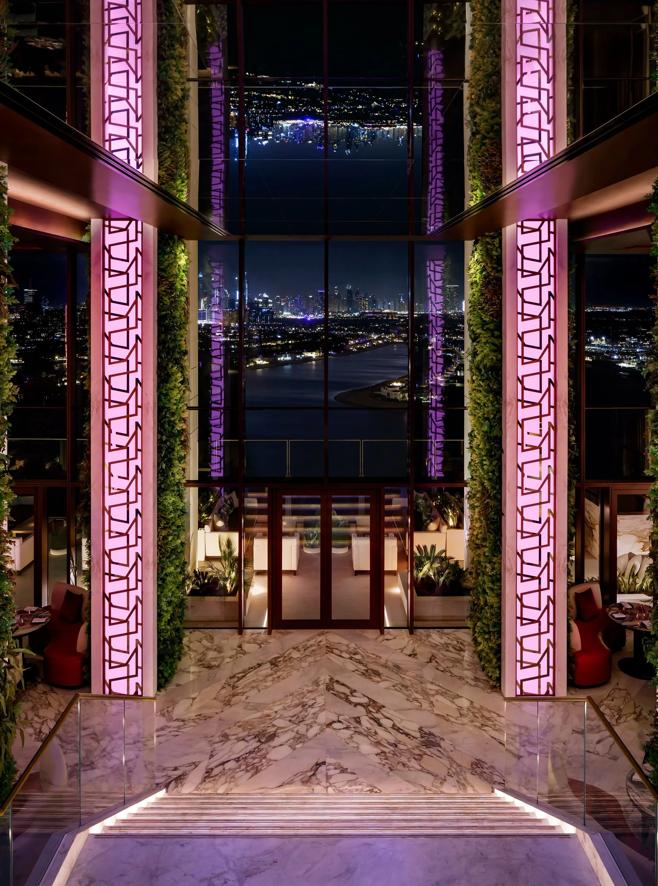 Elegant hotel lobby with marble flooring, illuminated pink geometric pillars, green vertical gardens, glass doors, and a view of a city skyline at night through large glass windows.