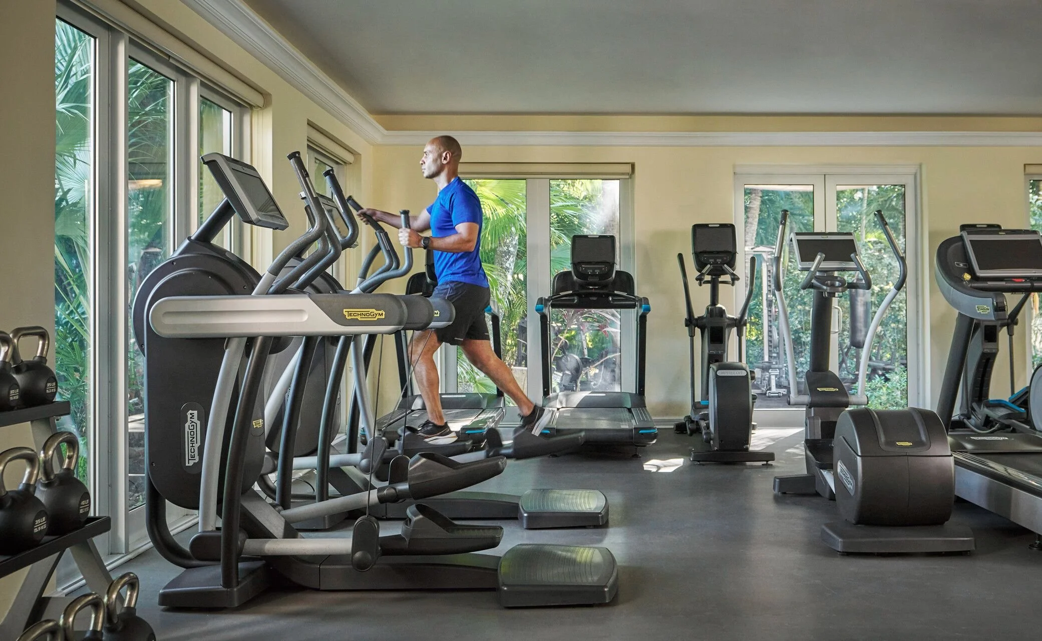 Man running on a treadmill in a gym with large windows and various exercise equipment.