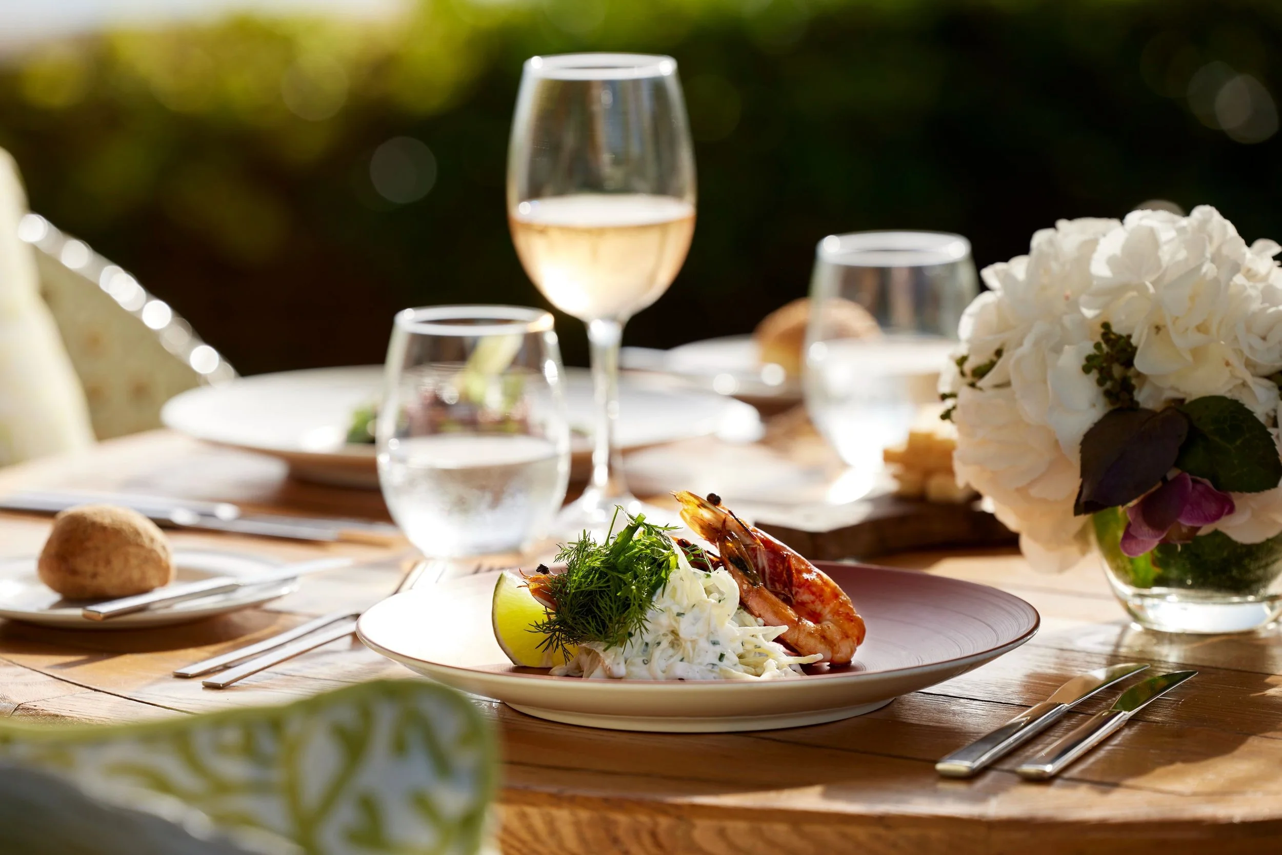 A plate with cooked shrimp garnished with herbs and lemon, set on a wooden dining table during a meal outdoors, with a glass of white wine, a floral arrangement, and other plates in the background.