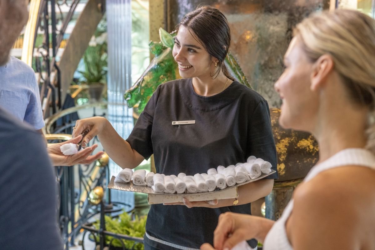 A young woman in a black shirt with a name tag, serving rolled towels to customers at a spa or wellness center, smiling indoors with plants and decorative elements in the background.