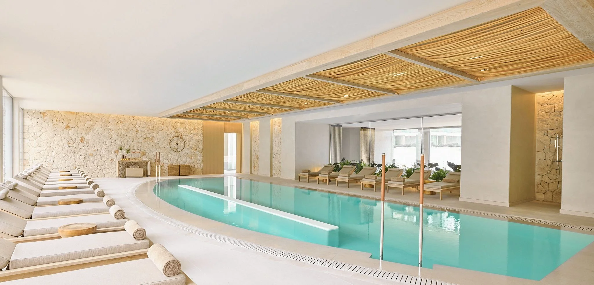 Indoor swimming pool with lounge chairs and wooden accents, stone wall, large windows, and modern decor.