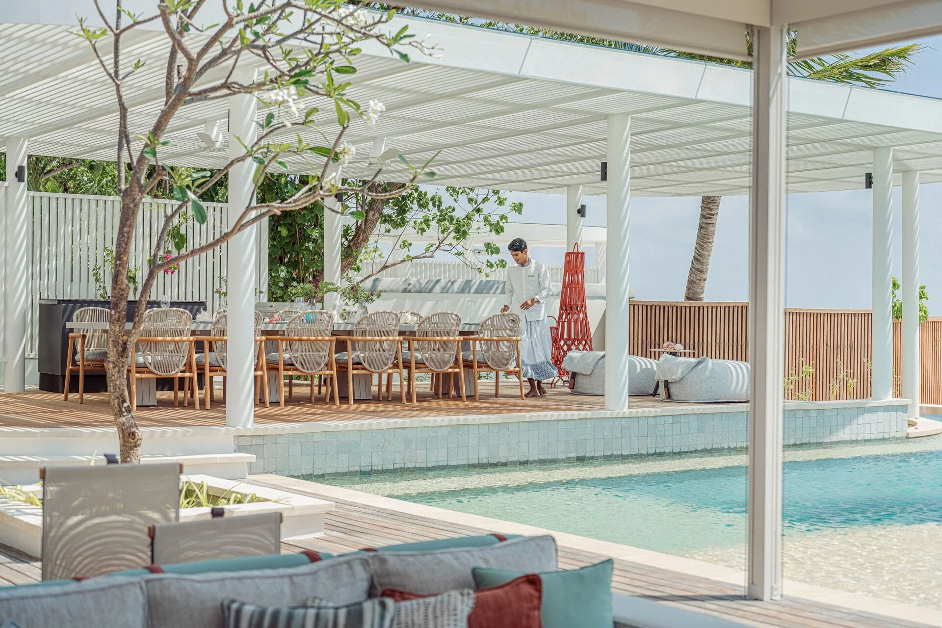 An outdoor patio area with a swimming pool surrounded by a deck and white furniture, including chairs and sofas. A person in white attire is standing near a long dining table with chairs, and tropical plants and trees are visible in the background under a white pergola.