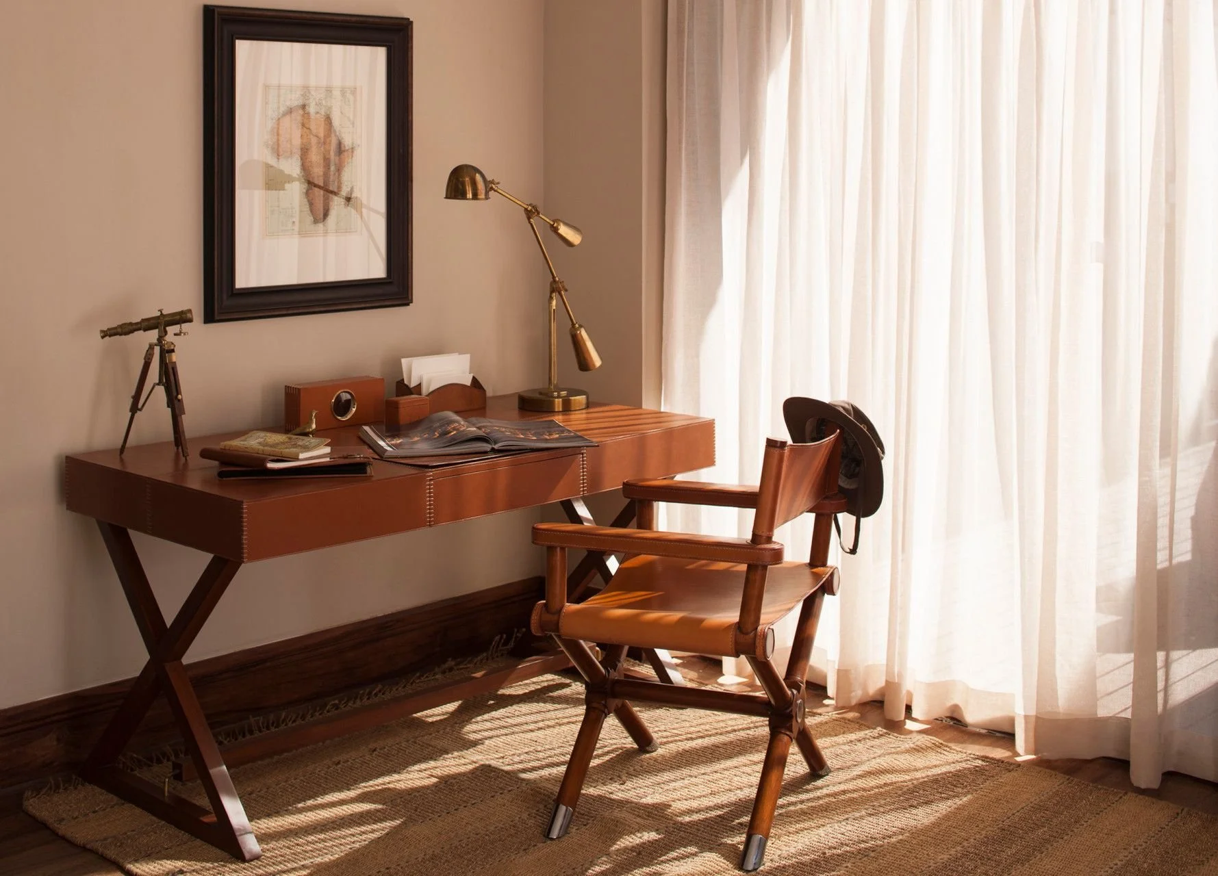 A home office corner with a wooden desk, a wooden chair with armrests, and a desk lamp. The desk has a telescope, some magazines, and a small box on it. Behind the desk is a framed map or artwork on the wall. To the right, there are white curtains covering a window, allowing sunlight to brighten the room.
