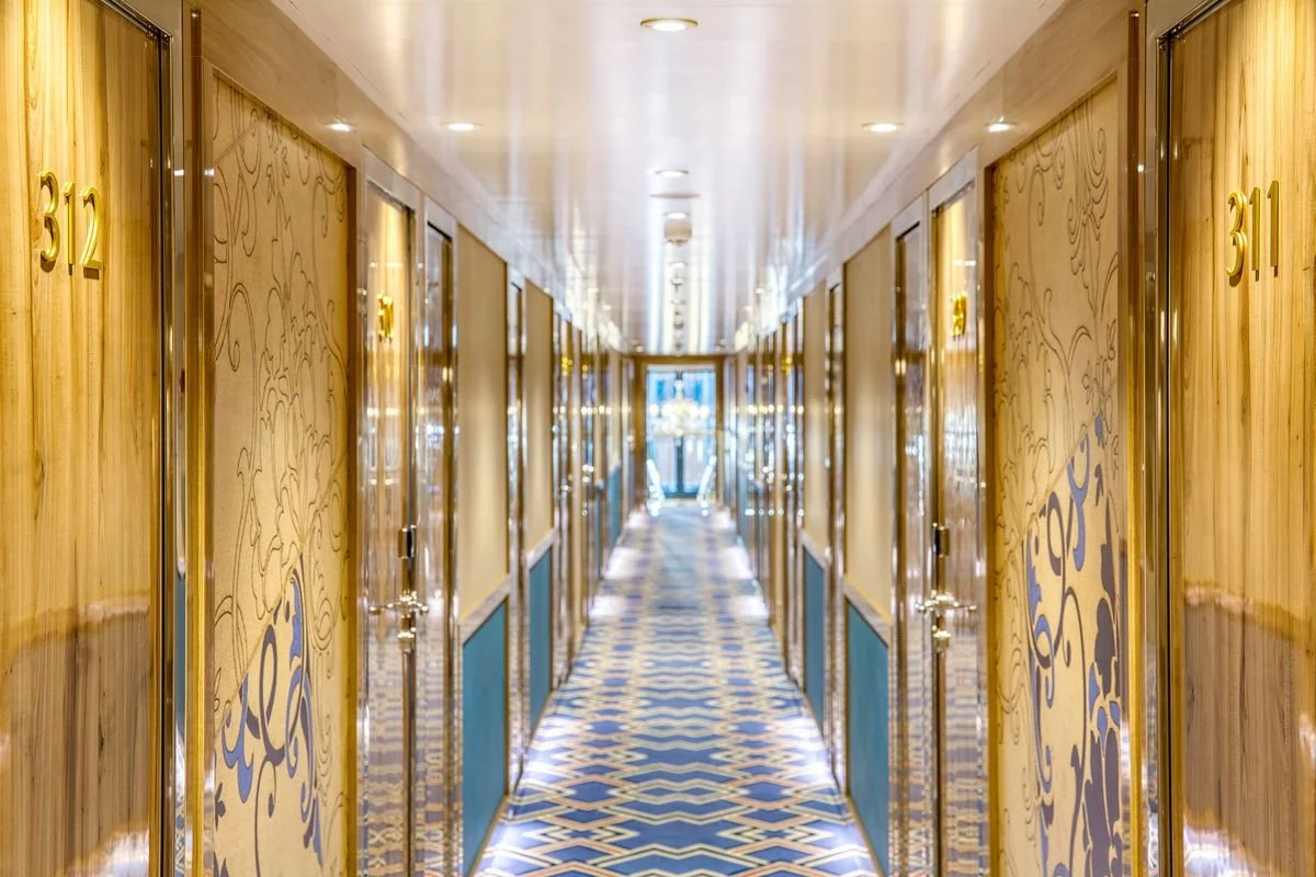 Hotel corridor with gold and blue patterned carpet, wood-paneled walls with decorative panels, gold numbers on the doors, and glass doors at the end of the hallway.