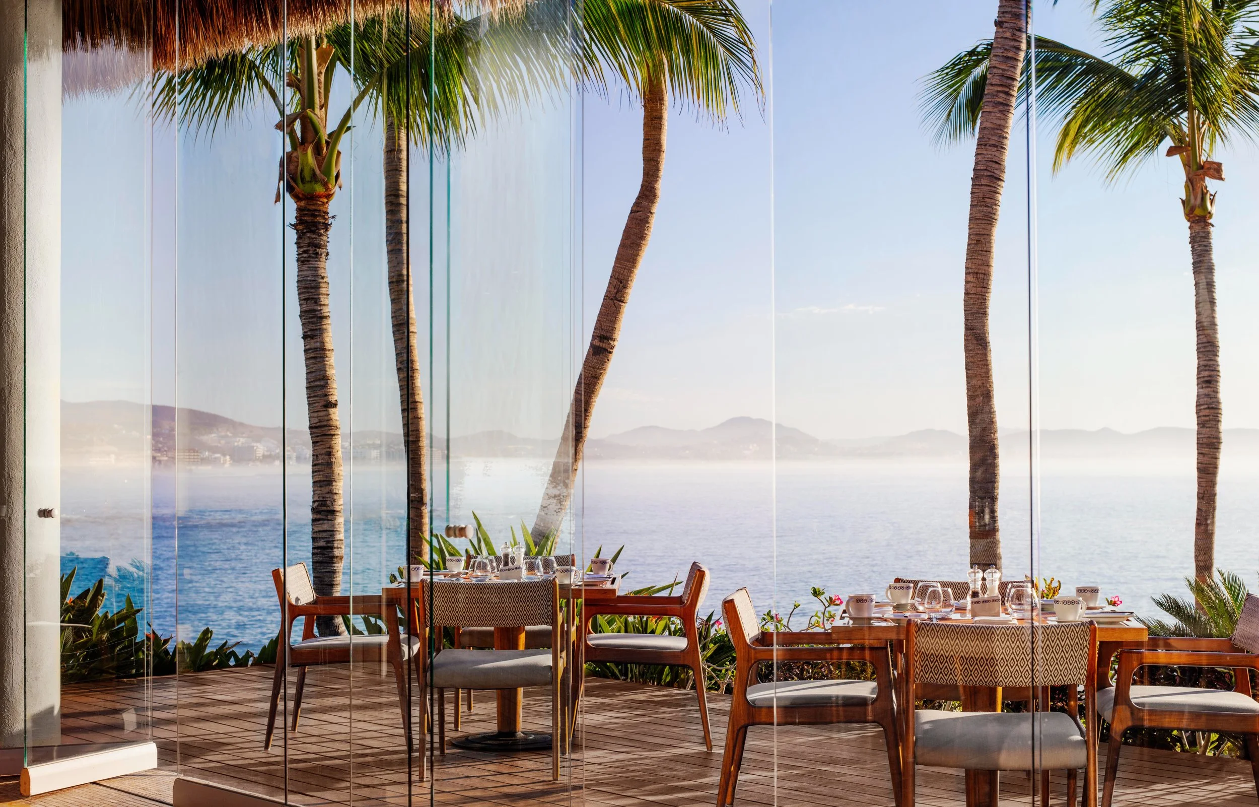 Dining area with wooden tables and chairs set with plates and glassware, overlooking a view of the ocean with palm trees and mountains in the background through glass walls.
