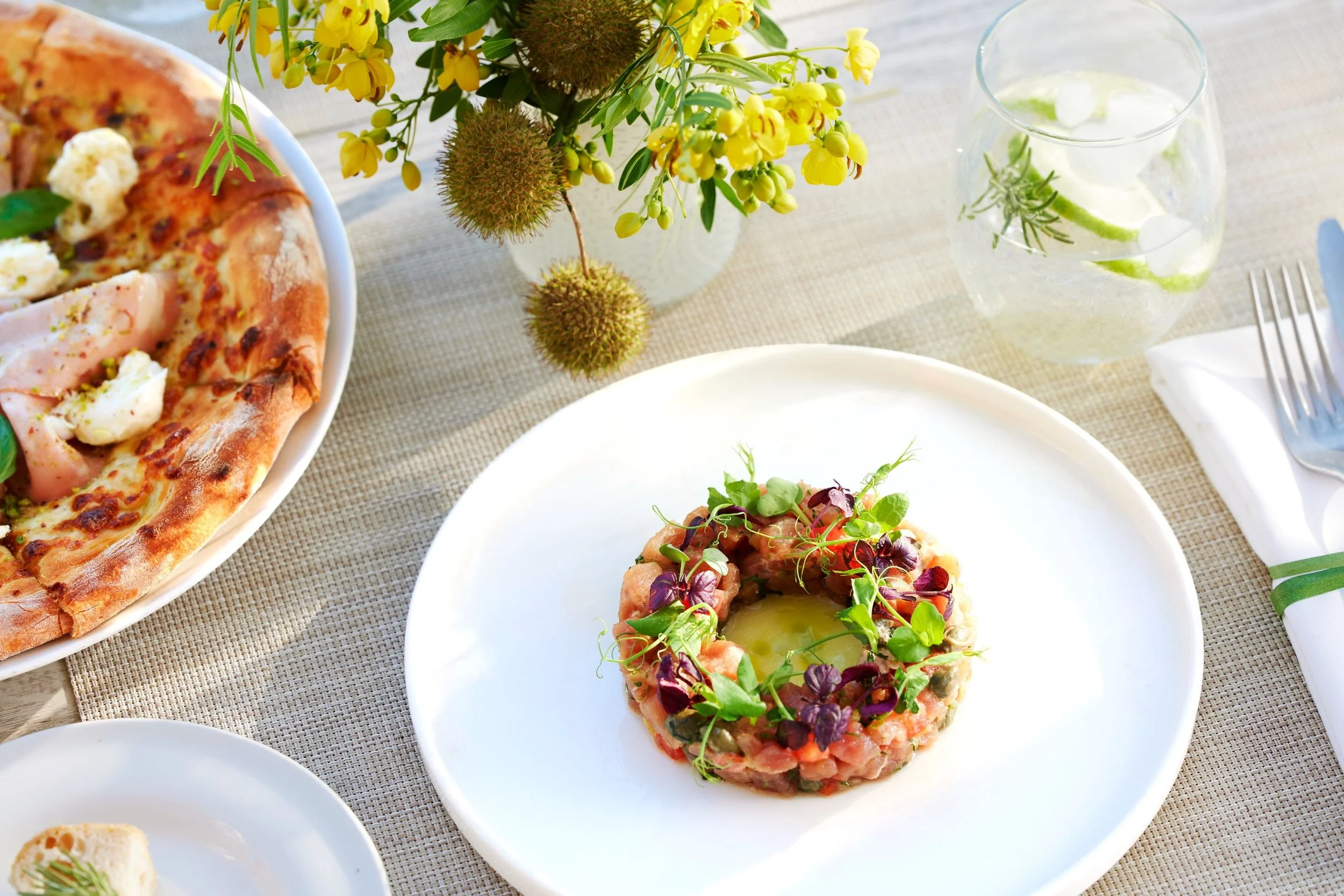 A plate of tuna tartare garnished with greens on a white plate, with a vase of yellow flowers and a glass of water with lime and herbs on a beige tablecloth.